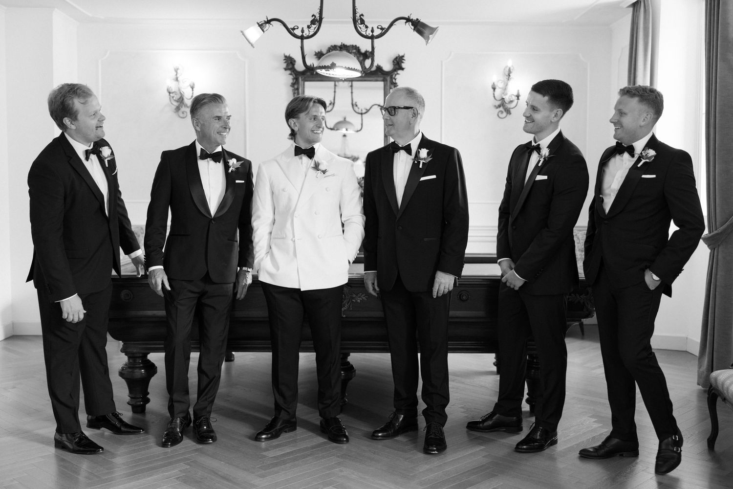 Six men in tuxedos and bow ties laughing together in an elegant room at a formal wedding celebration.