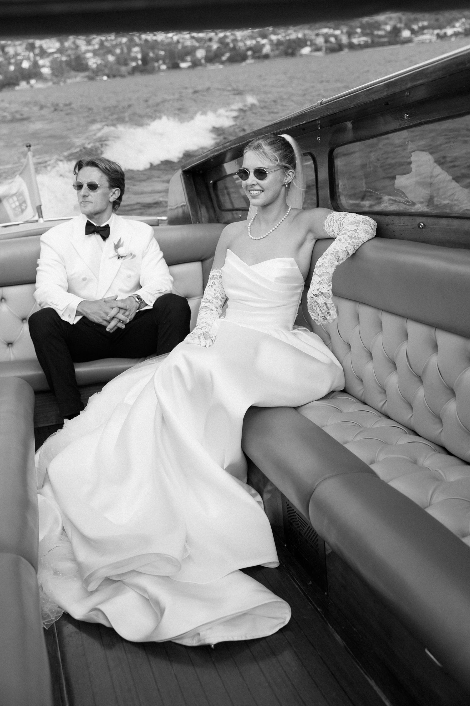 Bride and groom in sunglasses sit on tufted leather seats of a luxury boat, black and white wedding photo.