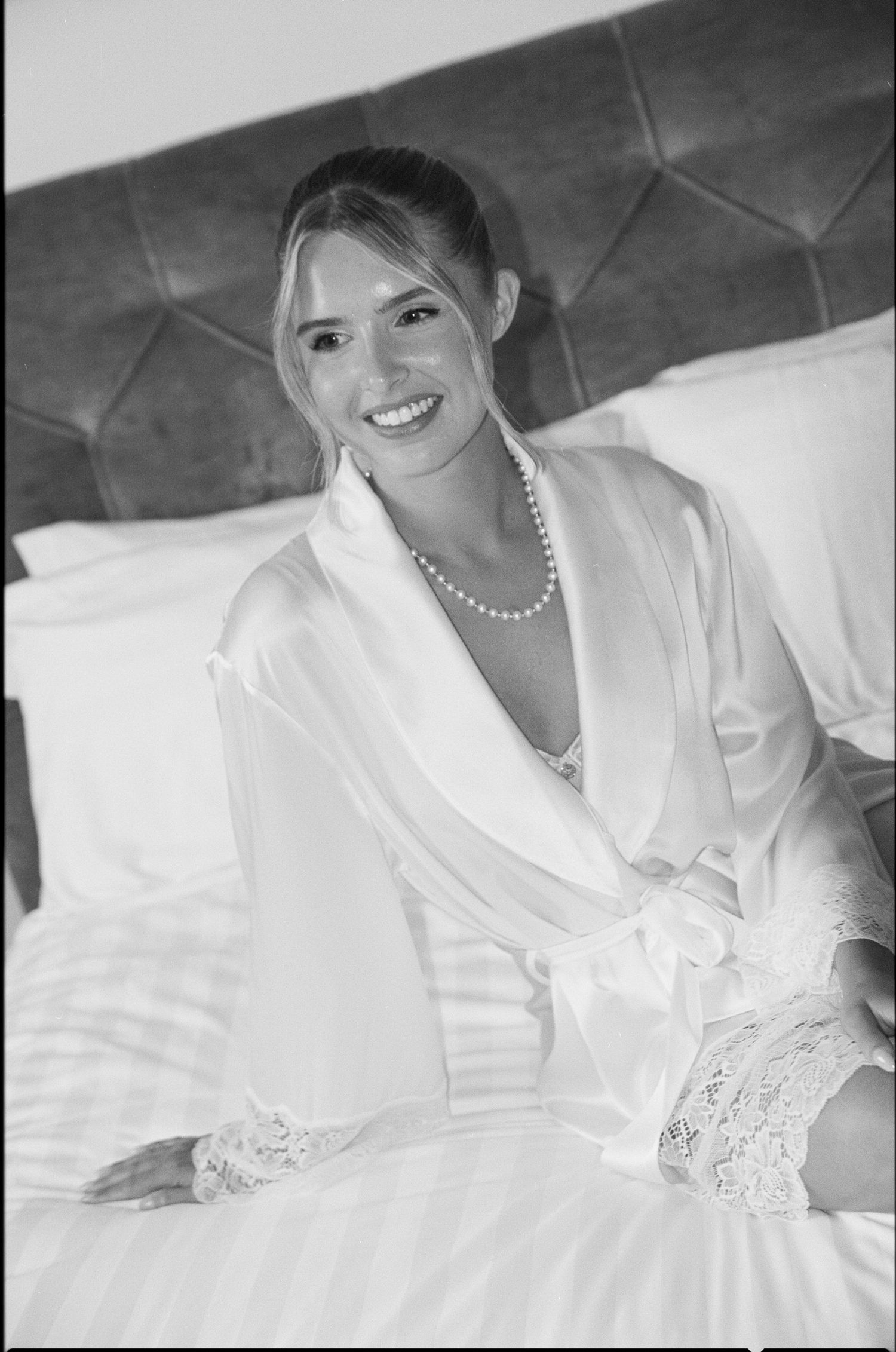Smiling bride in white satin robe and pearl necklace sitting on bed in black and white wedding photo.