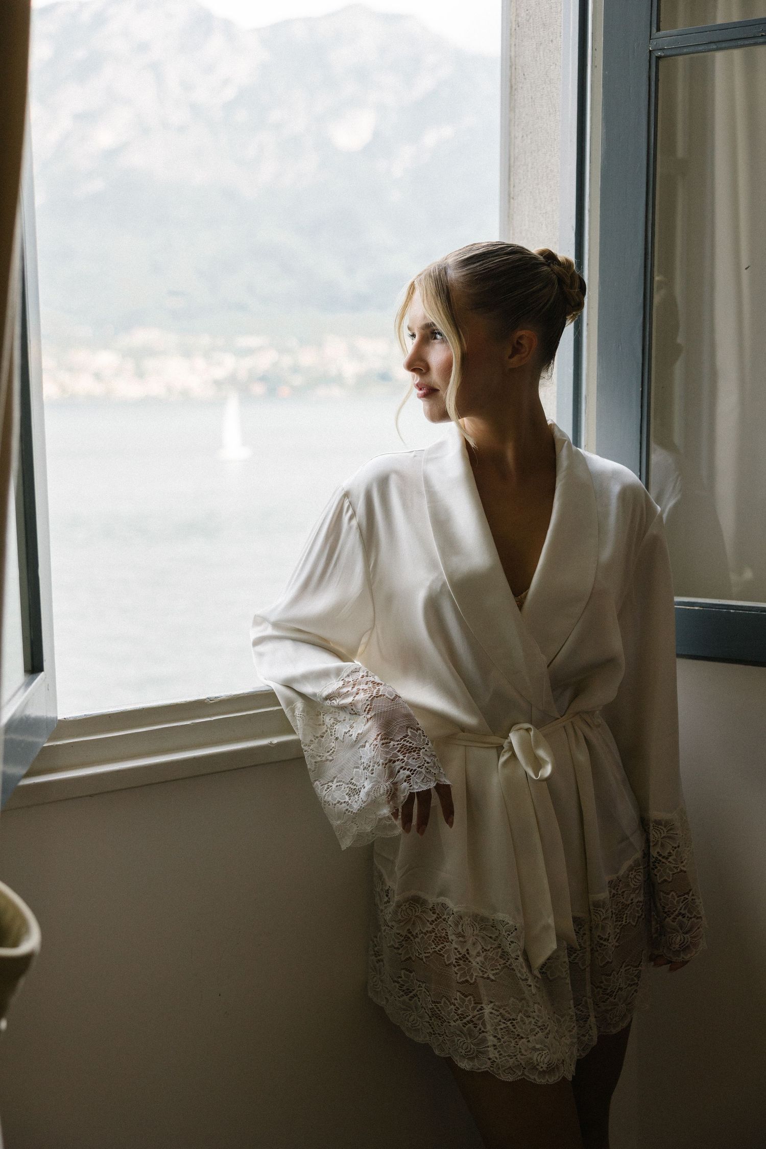 Bride in white lace robe gazing out window with mountain lake view on wedding morning.