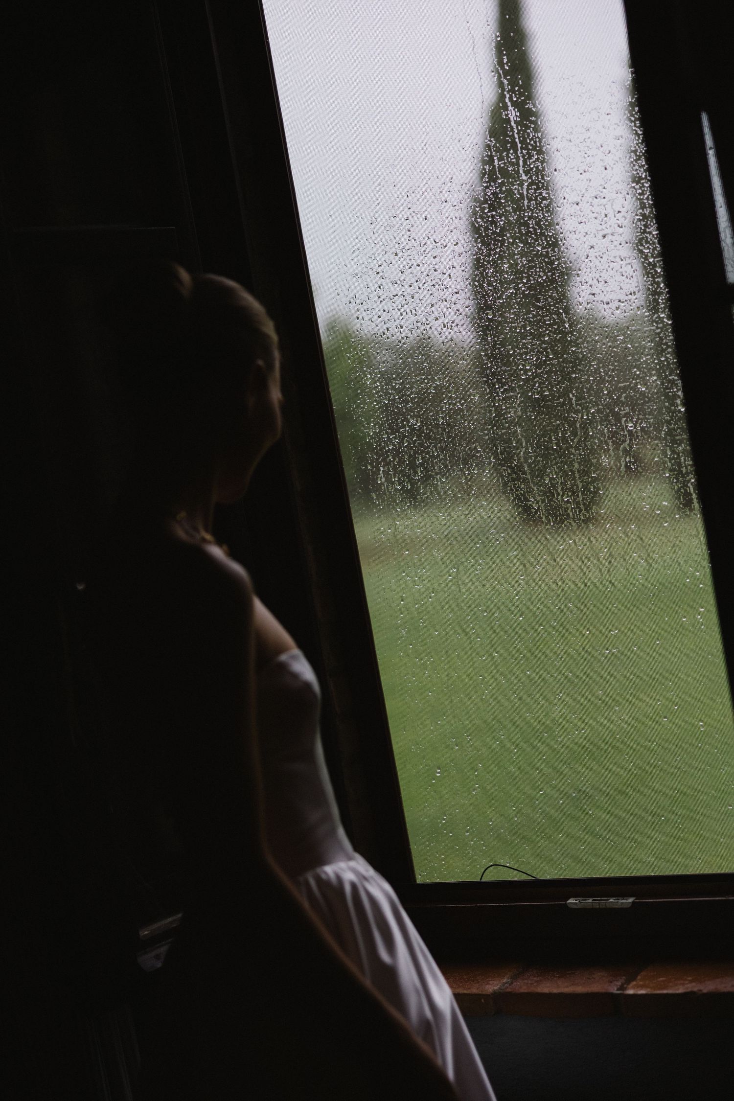Bride in white dress gazing through rain-covered window at lush green landscape on wedding day.