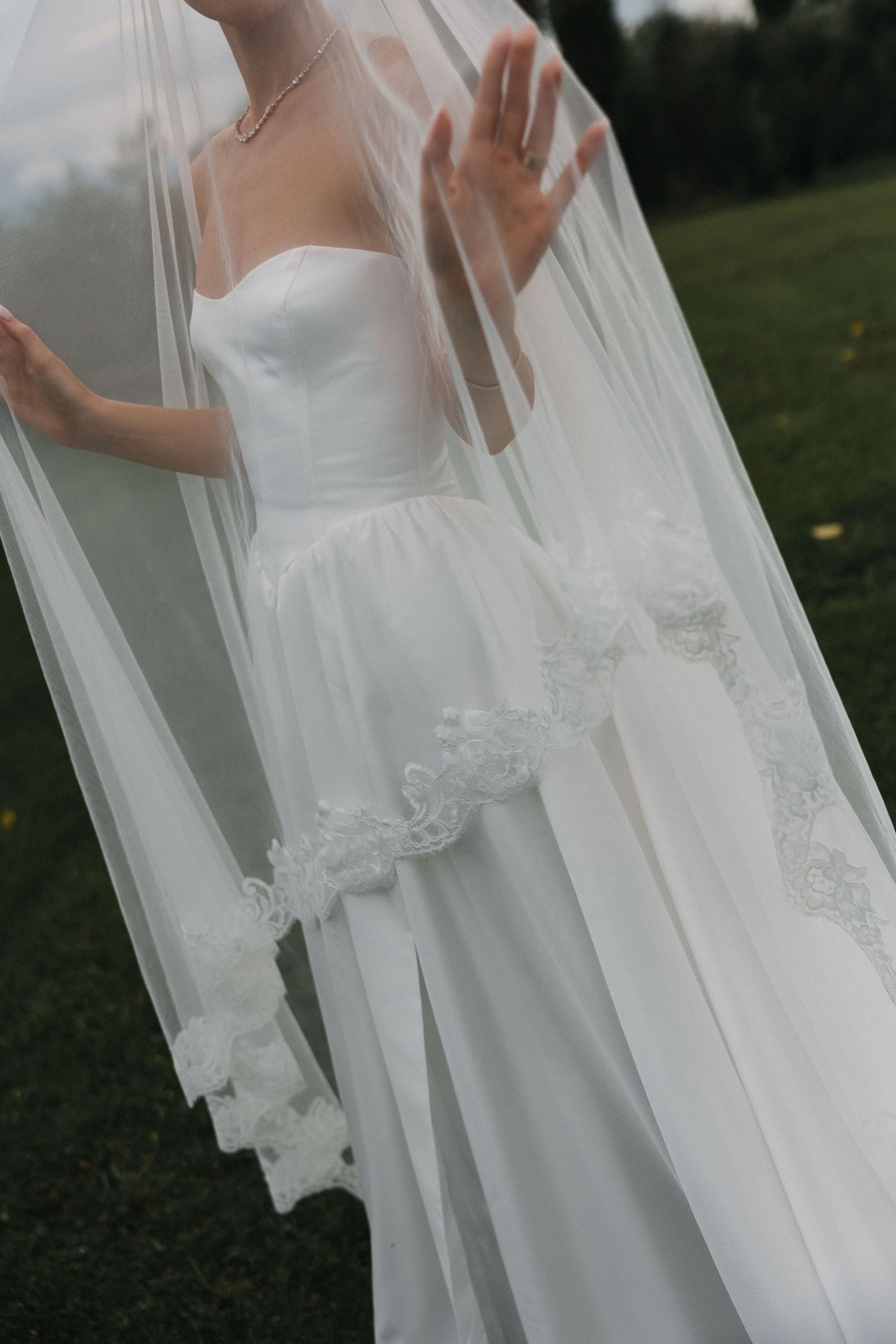 Bride in strapless white wedding gown with floral lace trim veil held up outdoors on green grass background.
