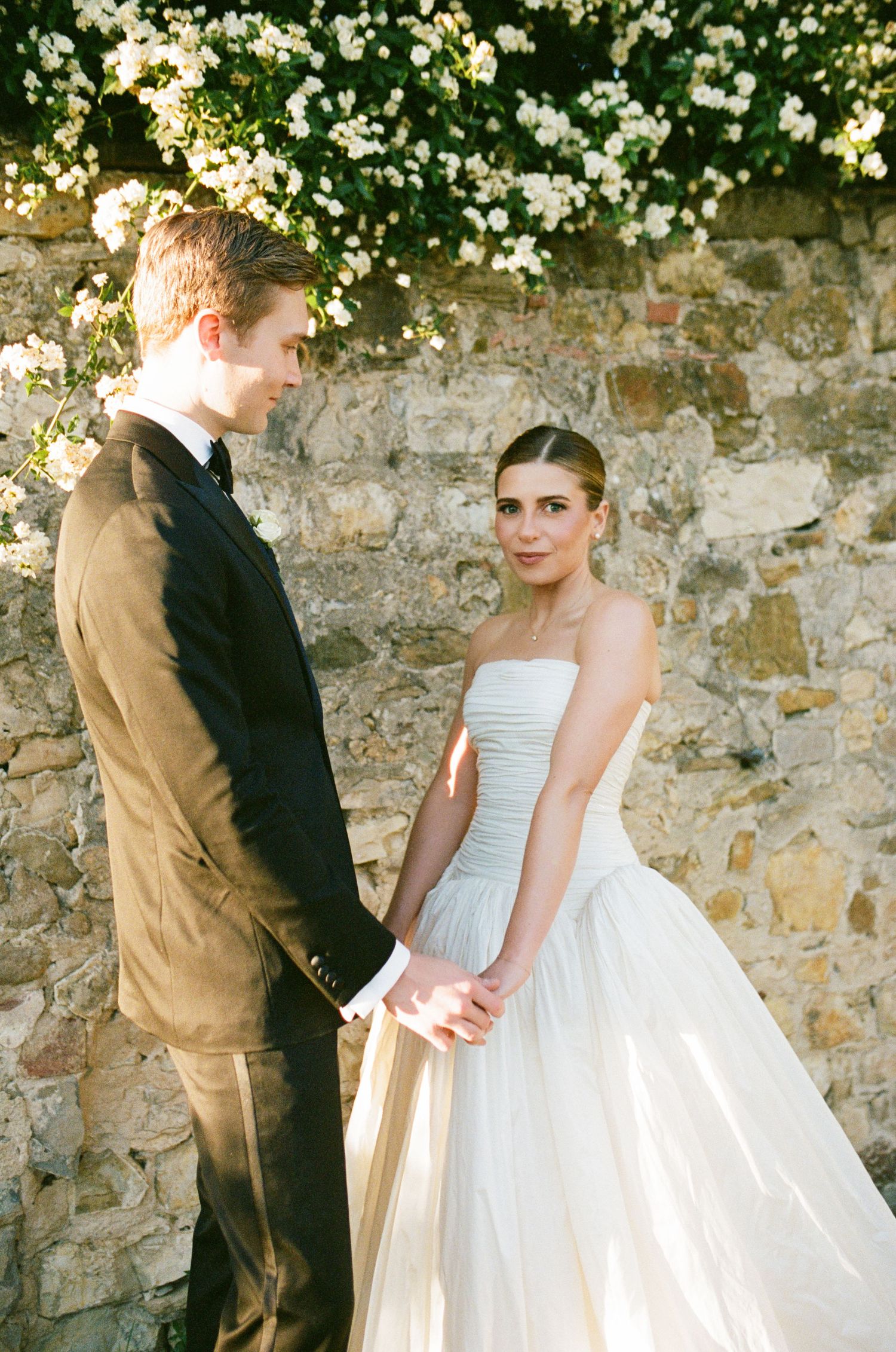 Bride in strapless white gown holds hands with groom in black tuxedo before a rustic stone wall with white flowers.