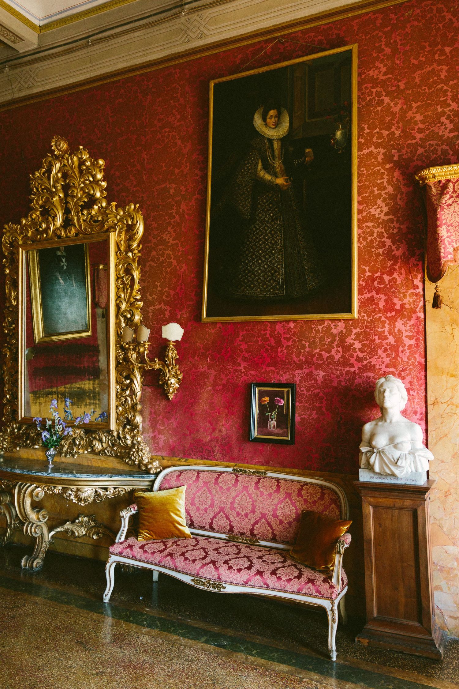 Ornate red damask room with gilded mirror, baroque settee, portrait painting, and marble bust sculpture.