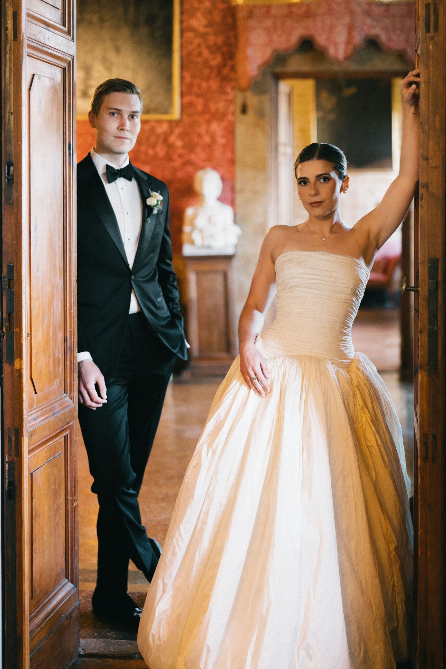 Bride in strapless ball gown and groom in black tuxedo posing in ornate doorway of elegant Italian villa.