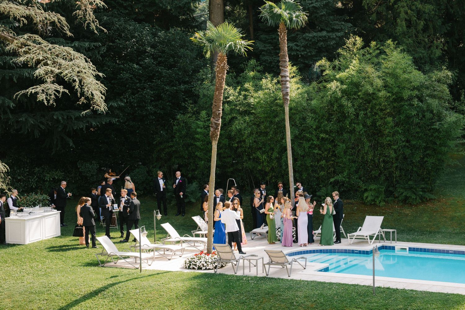 Elegant outdoor wedding reception by a pool surrounded by lush greenery with guests mingling on a sunny day.