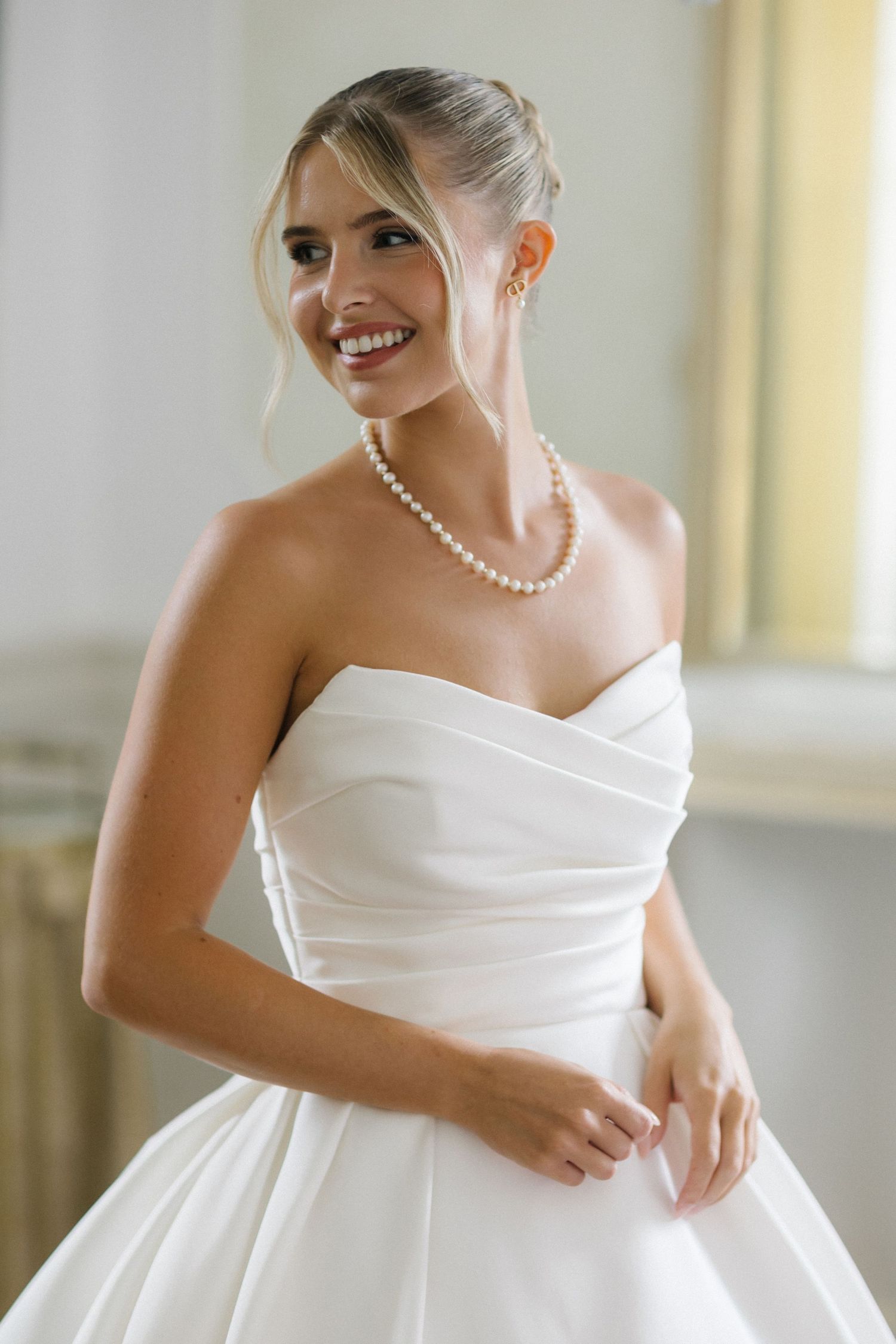 Smiling bride in strapless white ballgown wearing pearl necklace and earrings, posing elegantly in a bright room.