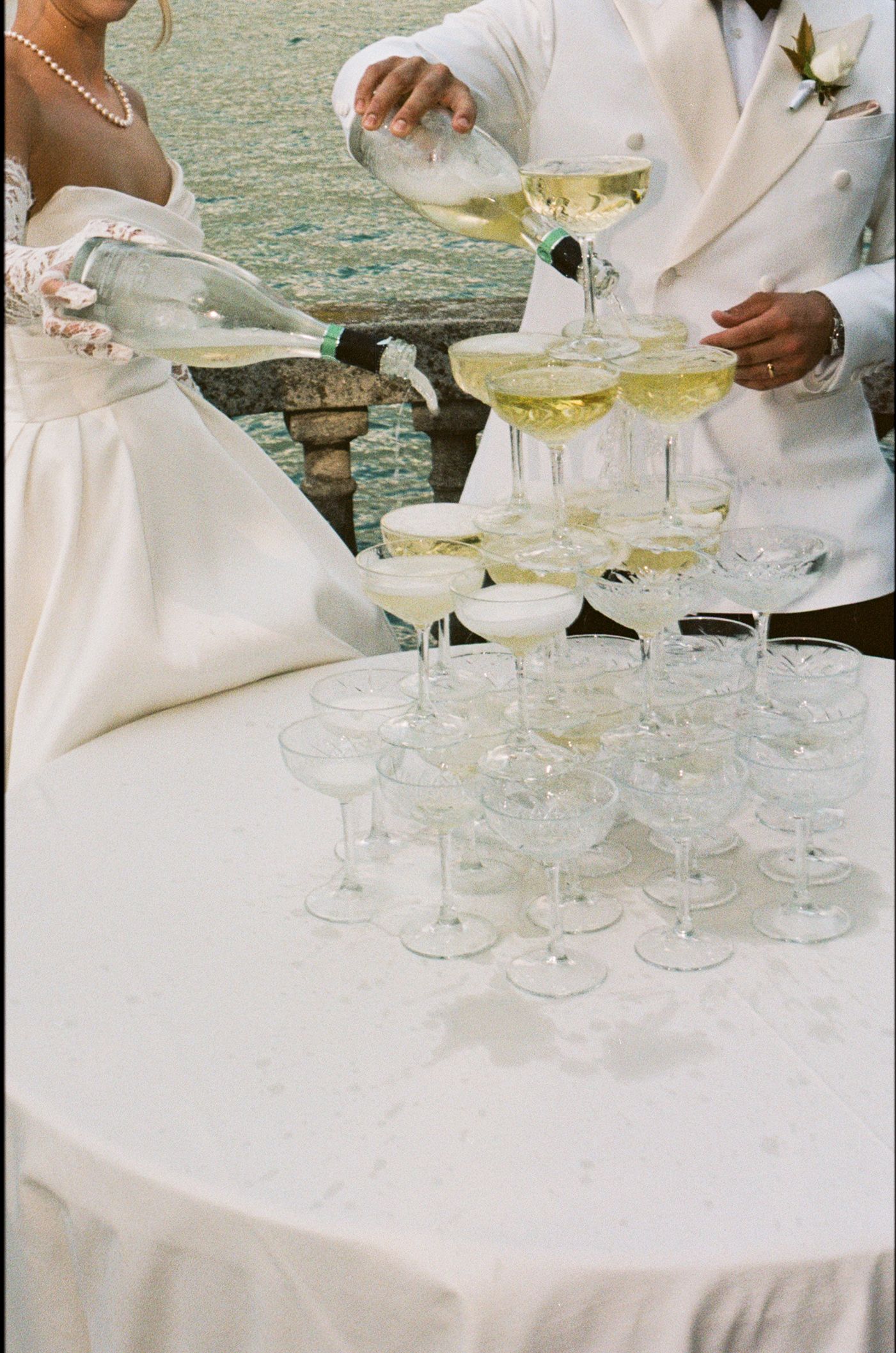 Bride and groom pouring champagne into a elegant tower of coupe glasses at a waterfront wedding reception.