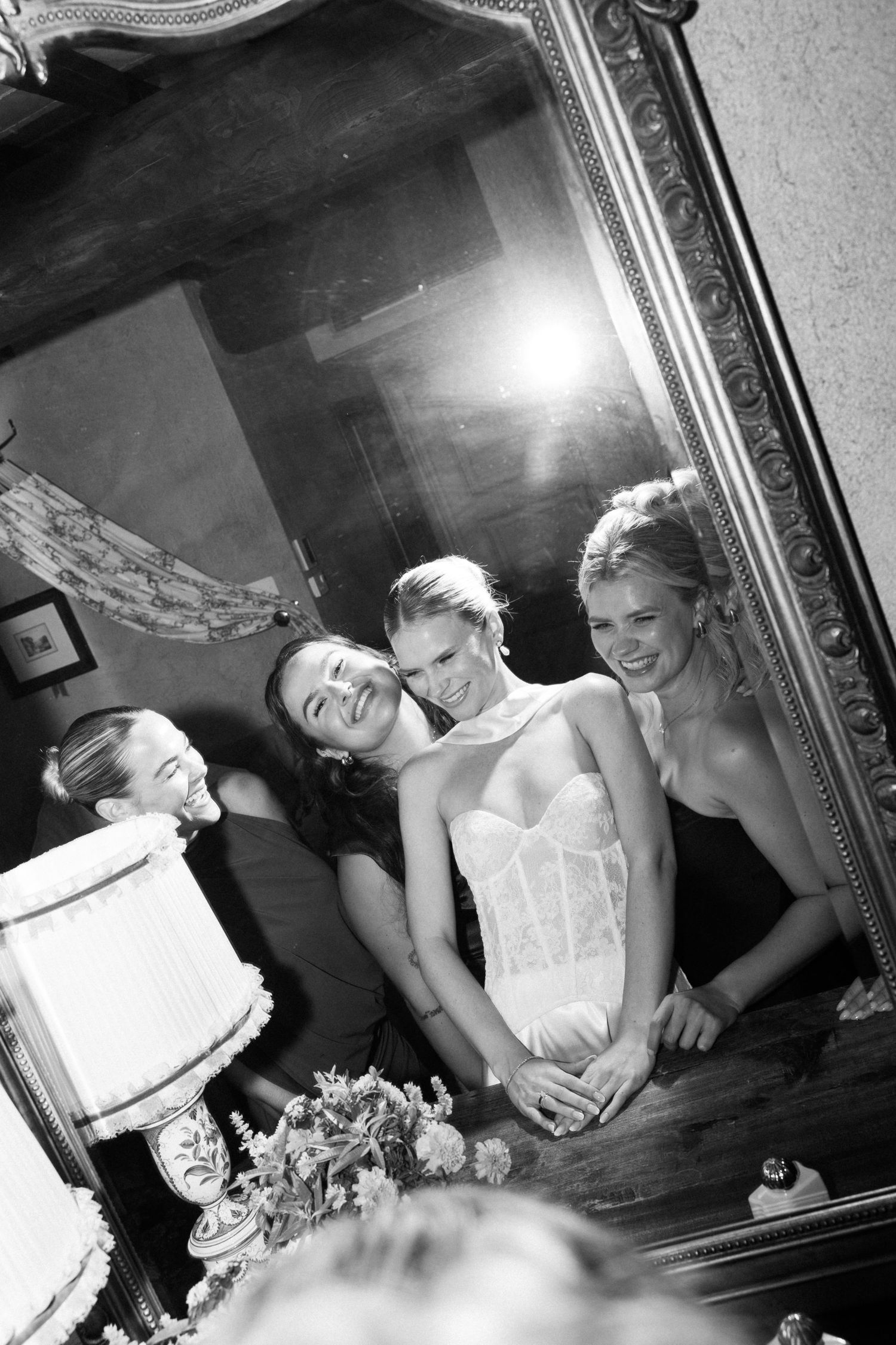 Black and white photo of a bride and bridesmaids smiling while cutting the wedding cake, reflected in an ornate gold mirror.