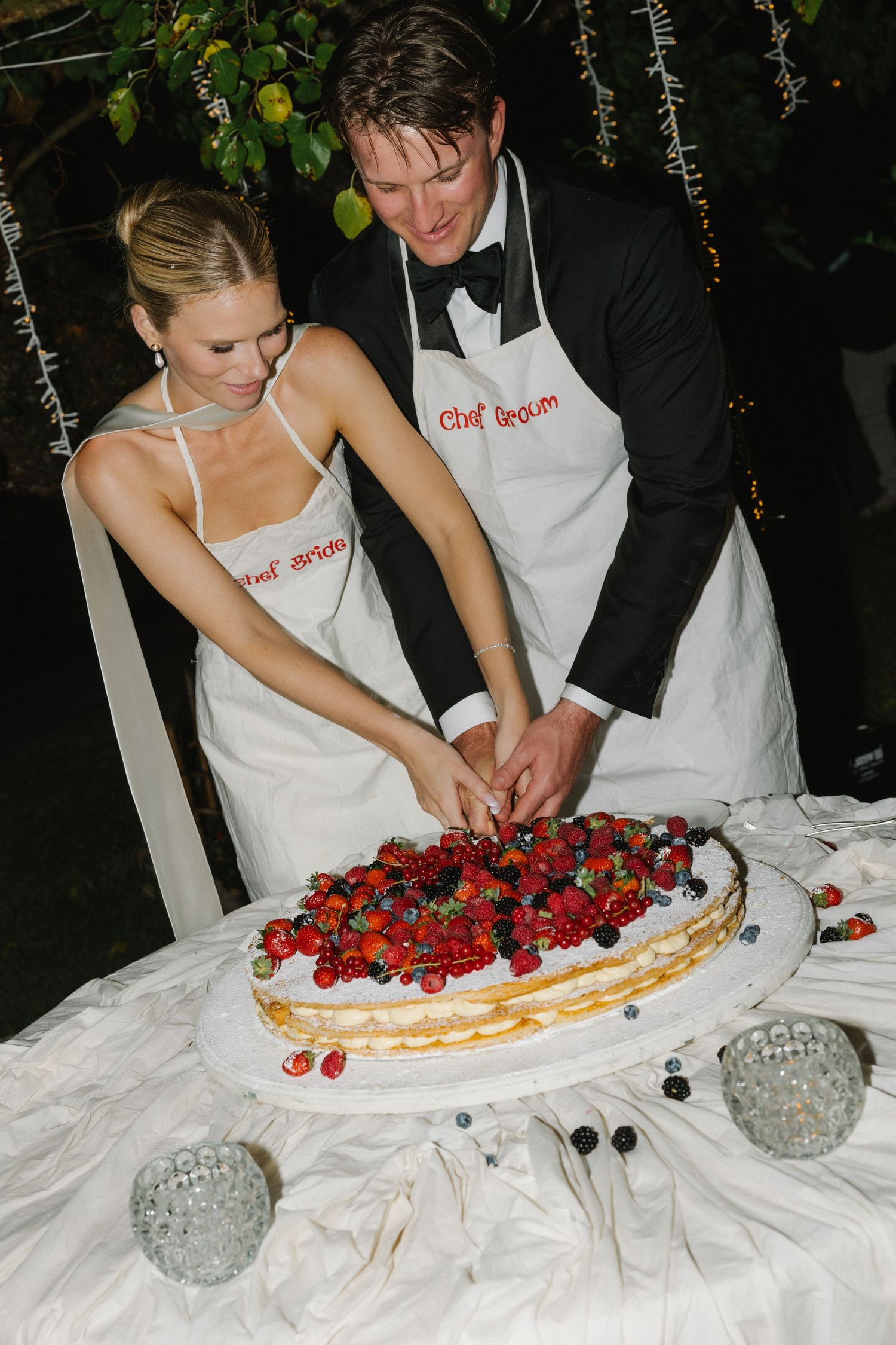 Bride and groom wearing chef aprons cutting a fresh berry wedding cake at an outdoor evening reception.