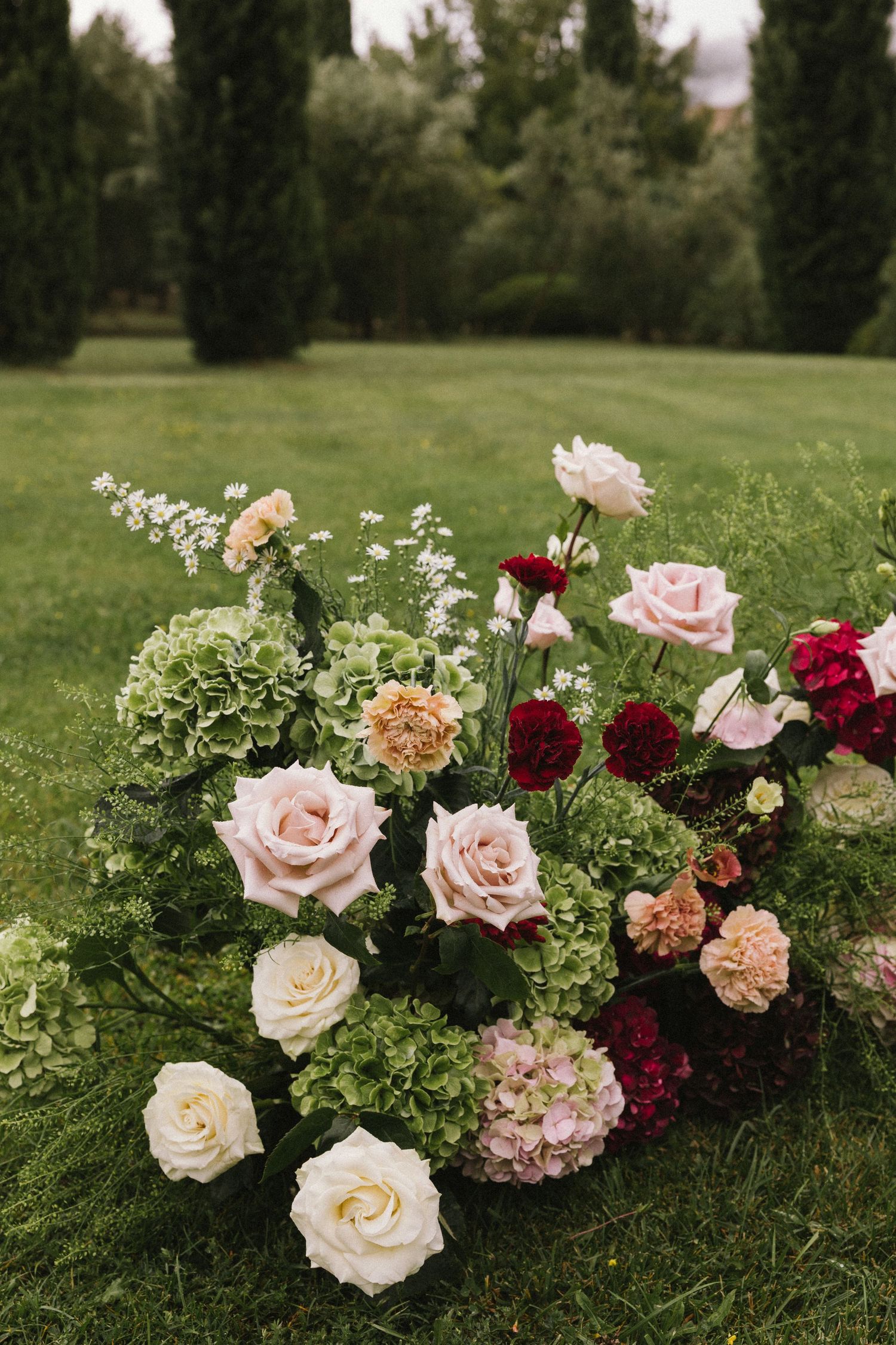 Lush floral arrangement with pink, cream, and burgundy roses and hydrangeas on green grass at an outdoor garden event.