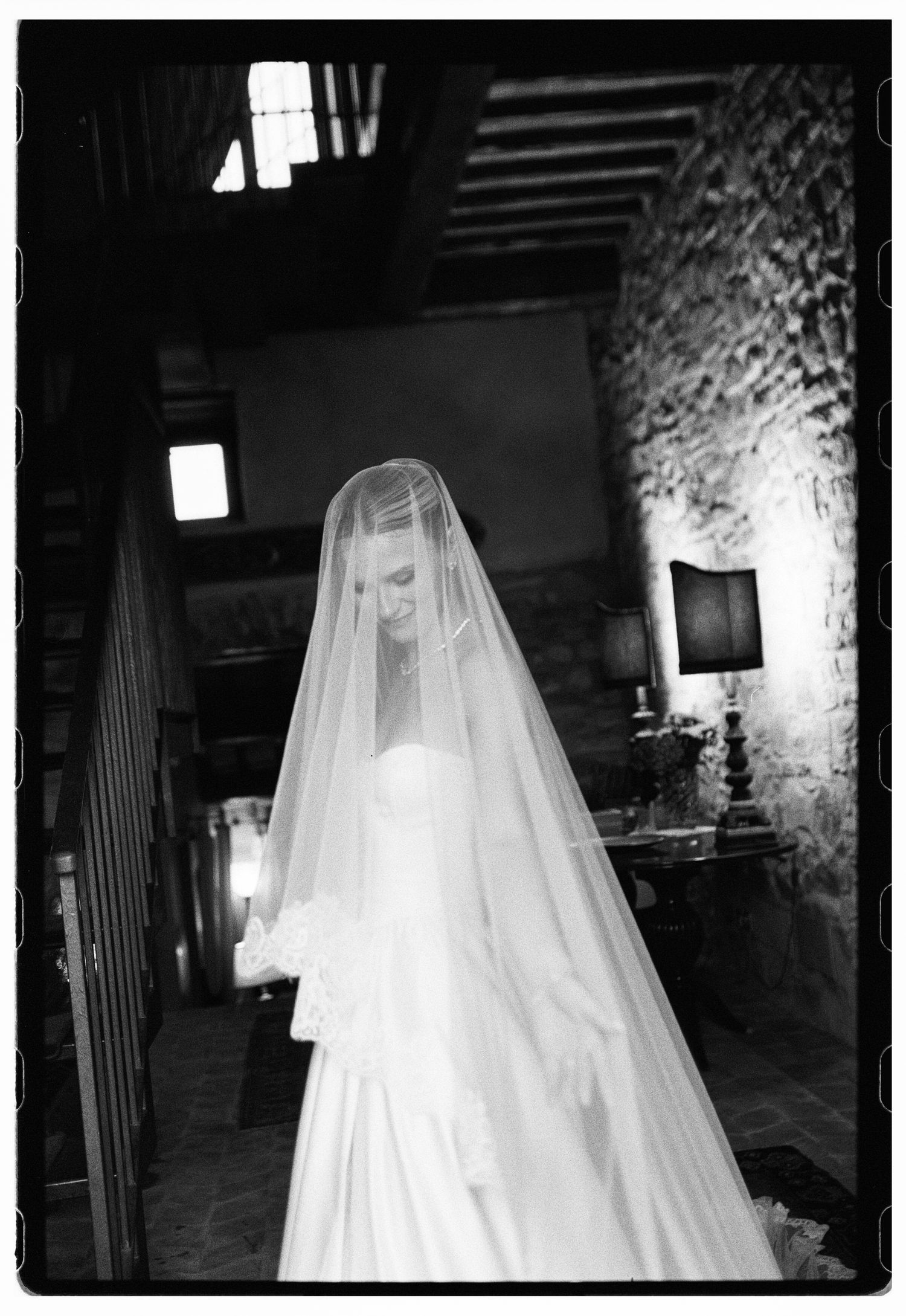Black and white photo of a bride in a flowing white gown and long veil standing in a rustic stone interior.