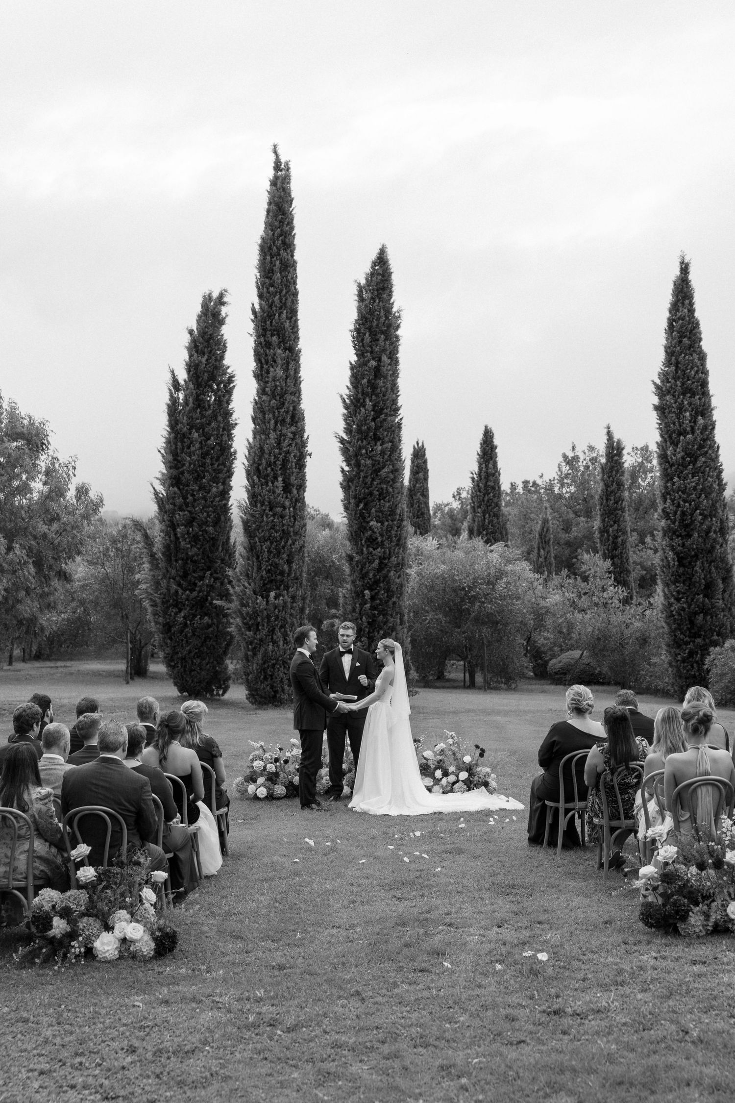 Black and white outdoor wedding ceremony with couple at altar, tall cypress trees, floral arrangements, and seated guests on a lush lawn.
