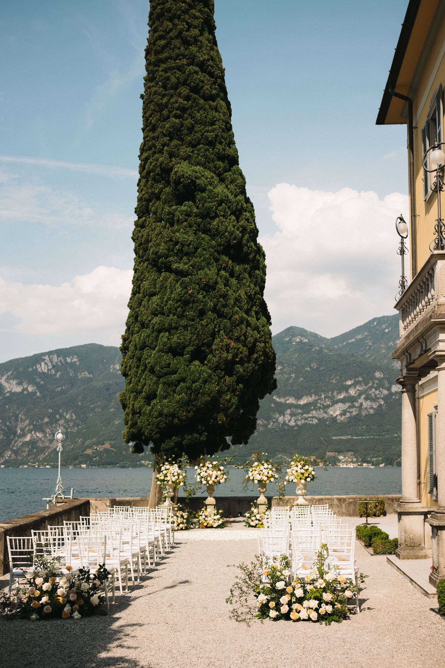 Elegant outdoor wedding ceremony setup with white chairs, floral arrangements, tall cypress tree, and stunning lake and mountain views.