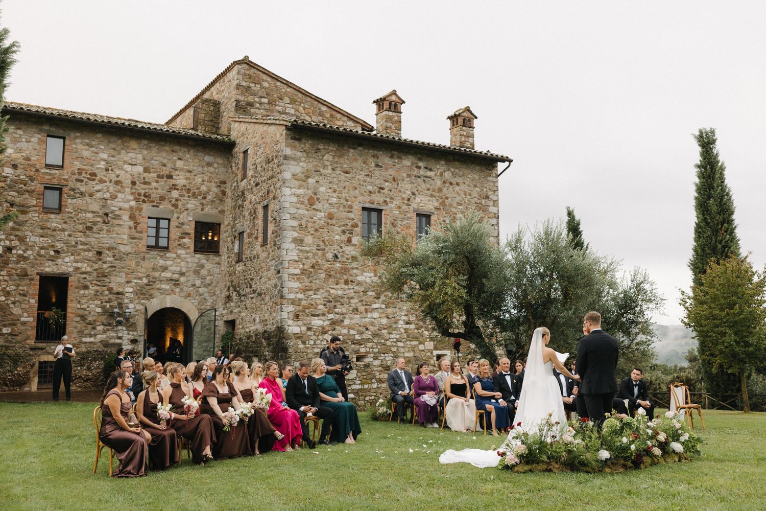 Outdoor wedding ceremony at a rustic Tuscan stone villa with guests seated on lawn chairs surrounded by lush greenery.