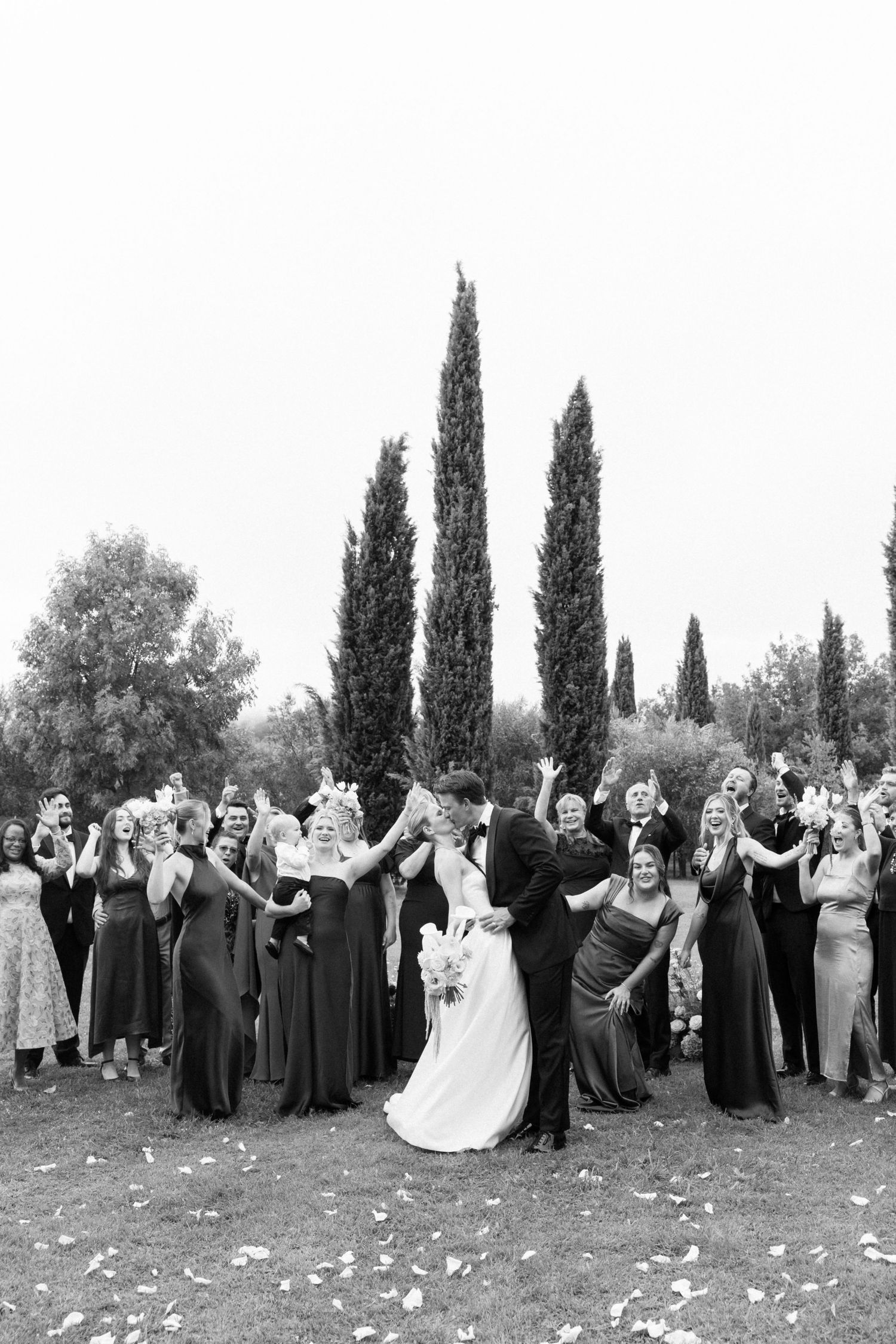 Black and white wedding photo of bride and groom kissing surrounded by cheering wedding party near tall cypress trees.