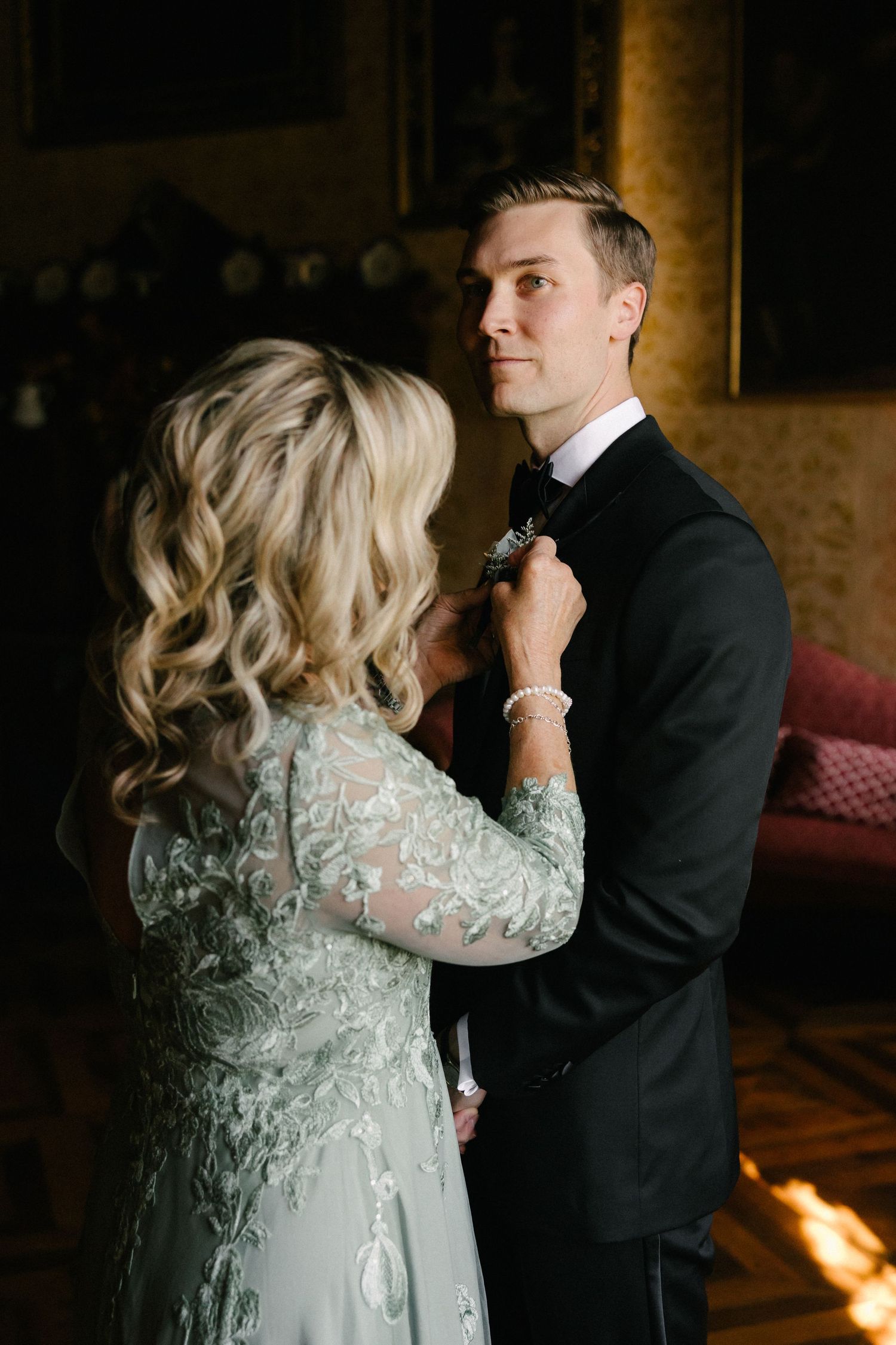 Blonde woman in lace gown adjusting groom's bow tie in elegant golden interior.