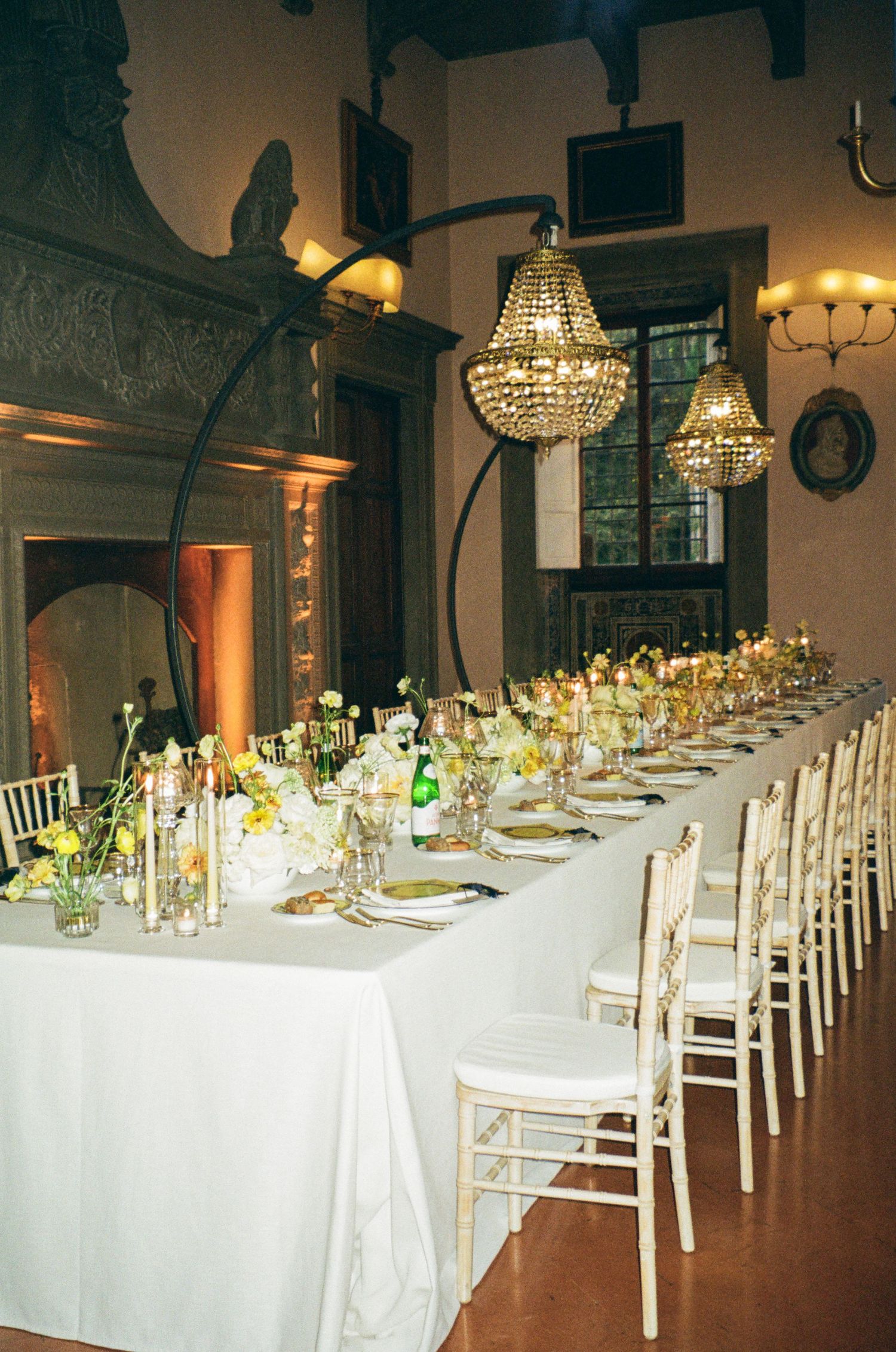 Elegant wedding reception with long white tablecloth, crystal chandeliers, white chiavari chairs, and floral centerpieces in a grand hall.