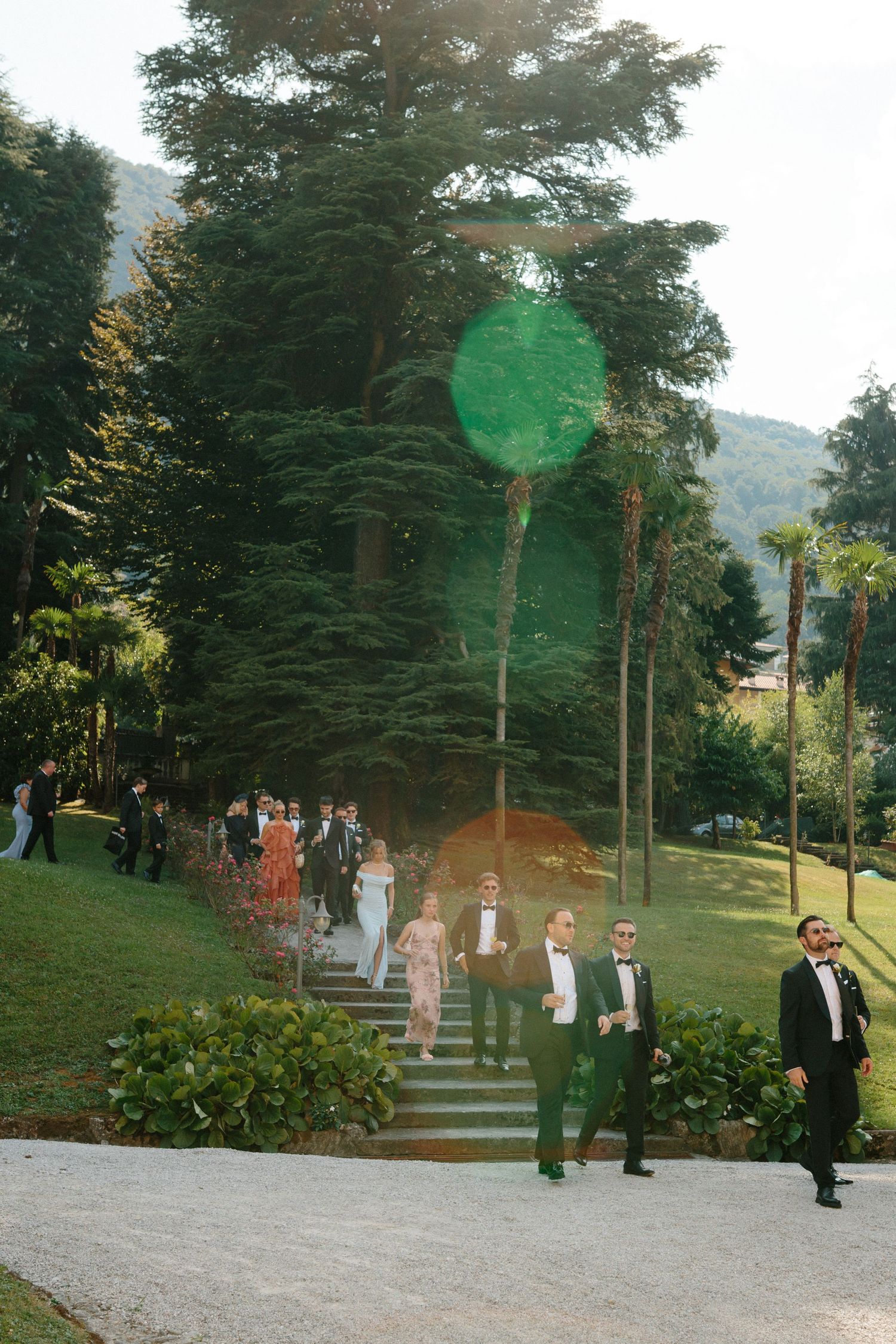 Wedding party descends stone steps in lush garden, men in green tuxedos, surrounded by tall trees and mountains.