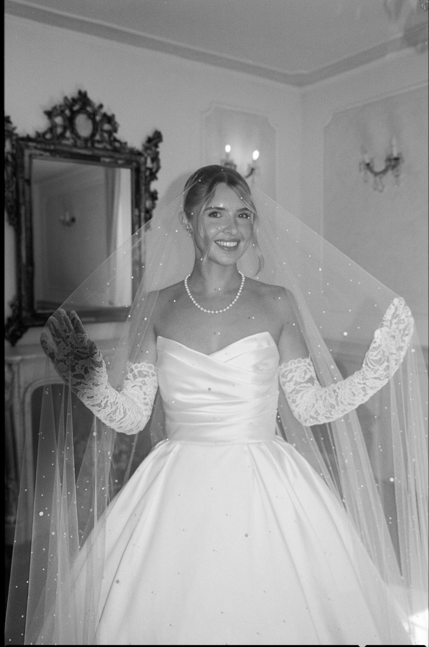 Smiling bride in strapless satin gown, lace gloves, pearl necklace, and veil in elegant black and white photo.