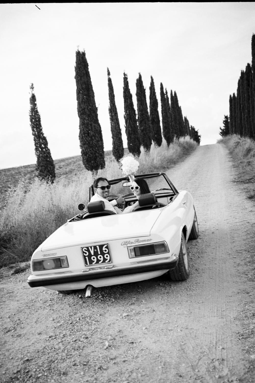Couple in white convertible car on rural Tuscan dirt road lined with cypress trees, black and white photo.