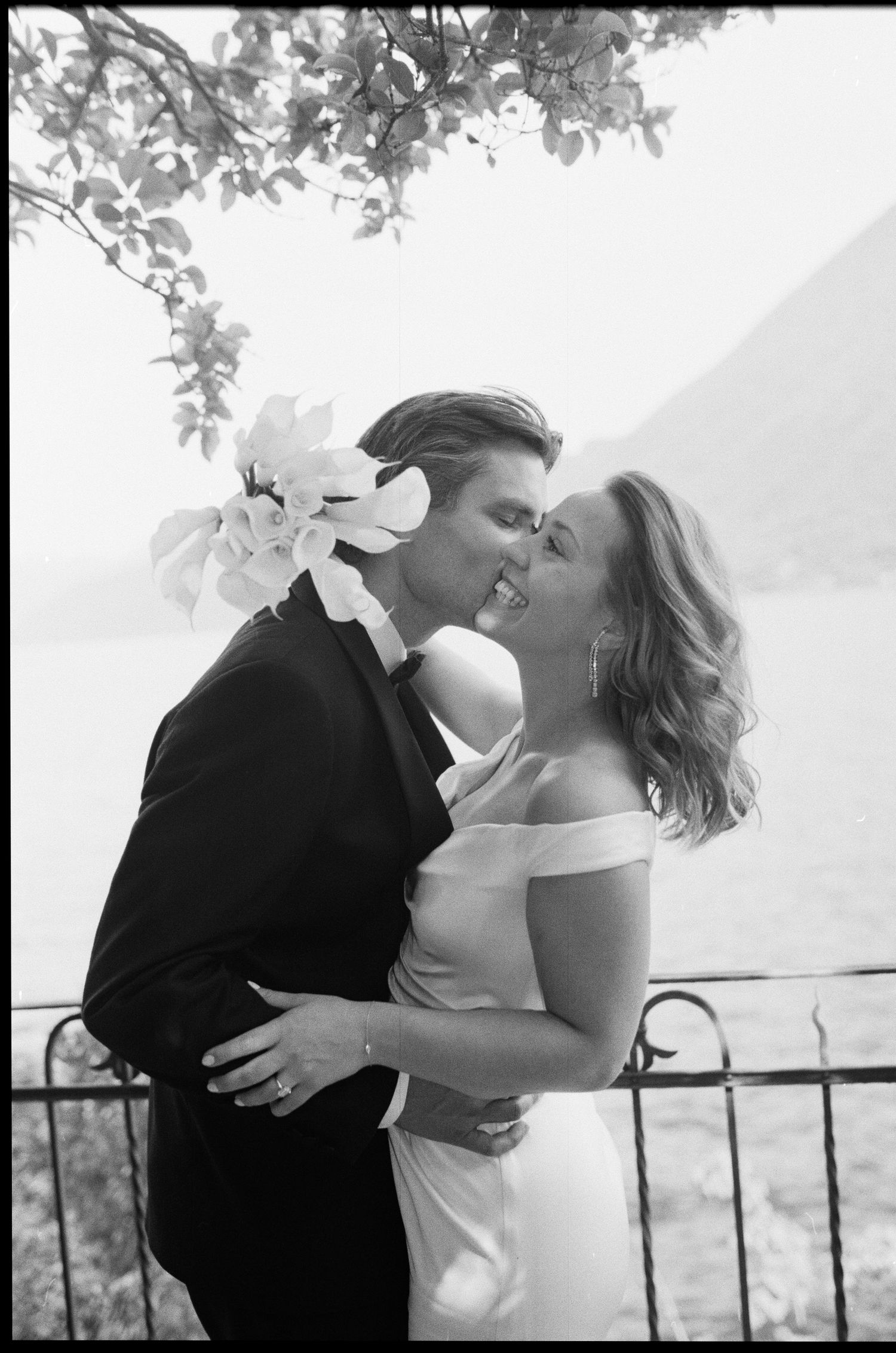 Black and white romantic photo of bride and groom kissing by a lakeside railing.