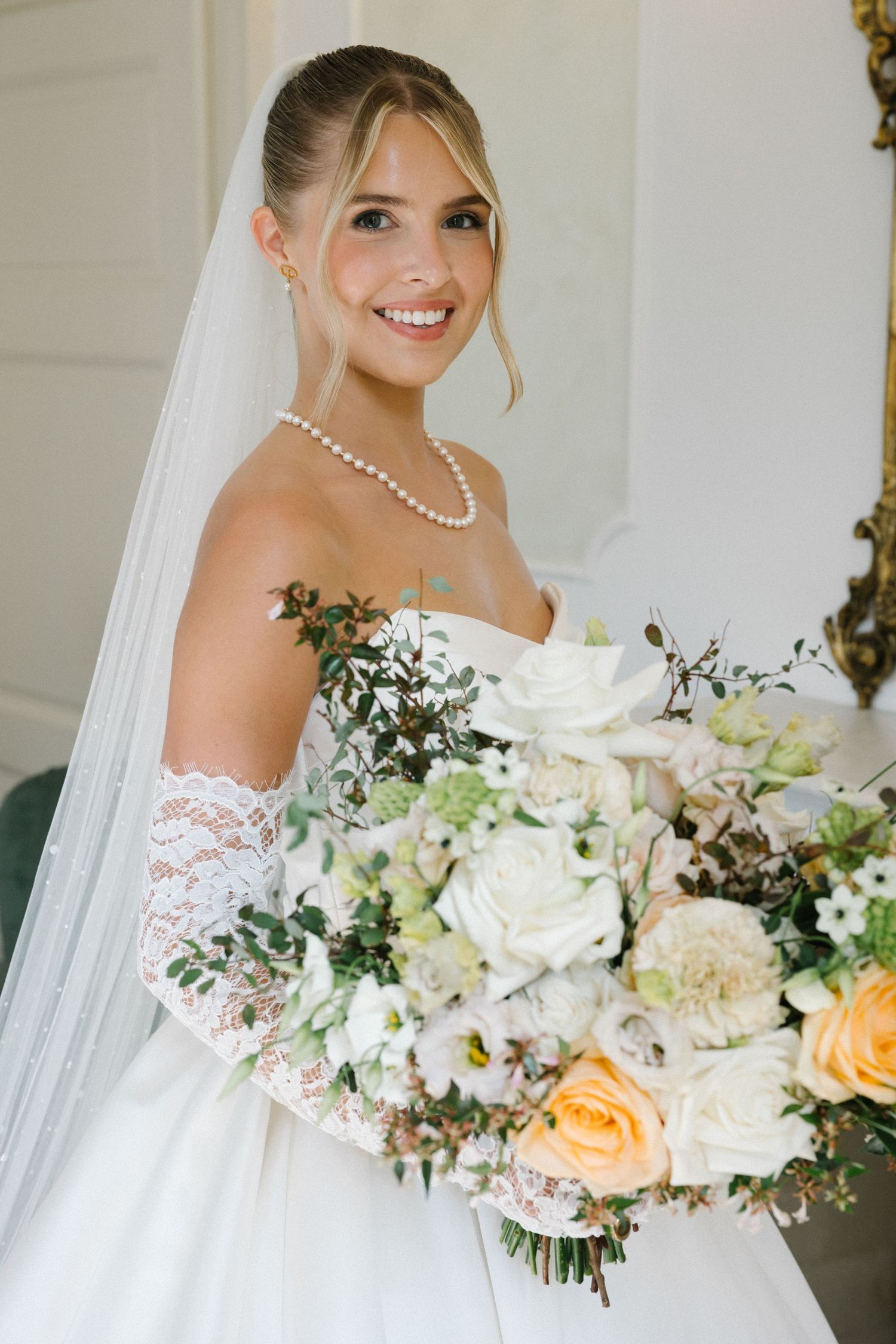 Smiling bride in lace off-shoulder gown, pearl necklace, and veil holds large white and peach floral bouquet.
