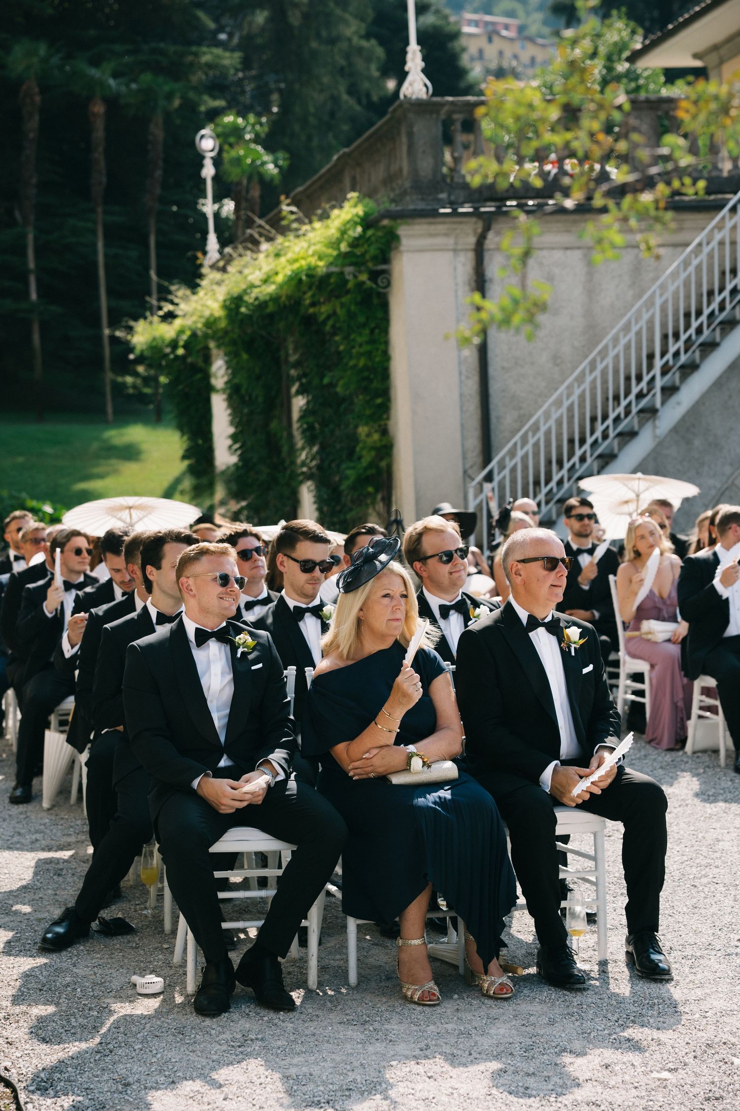 Elegantly dressed wedding guests in black tie attire seated outdoors at a sunlit garden ceremony.