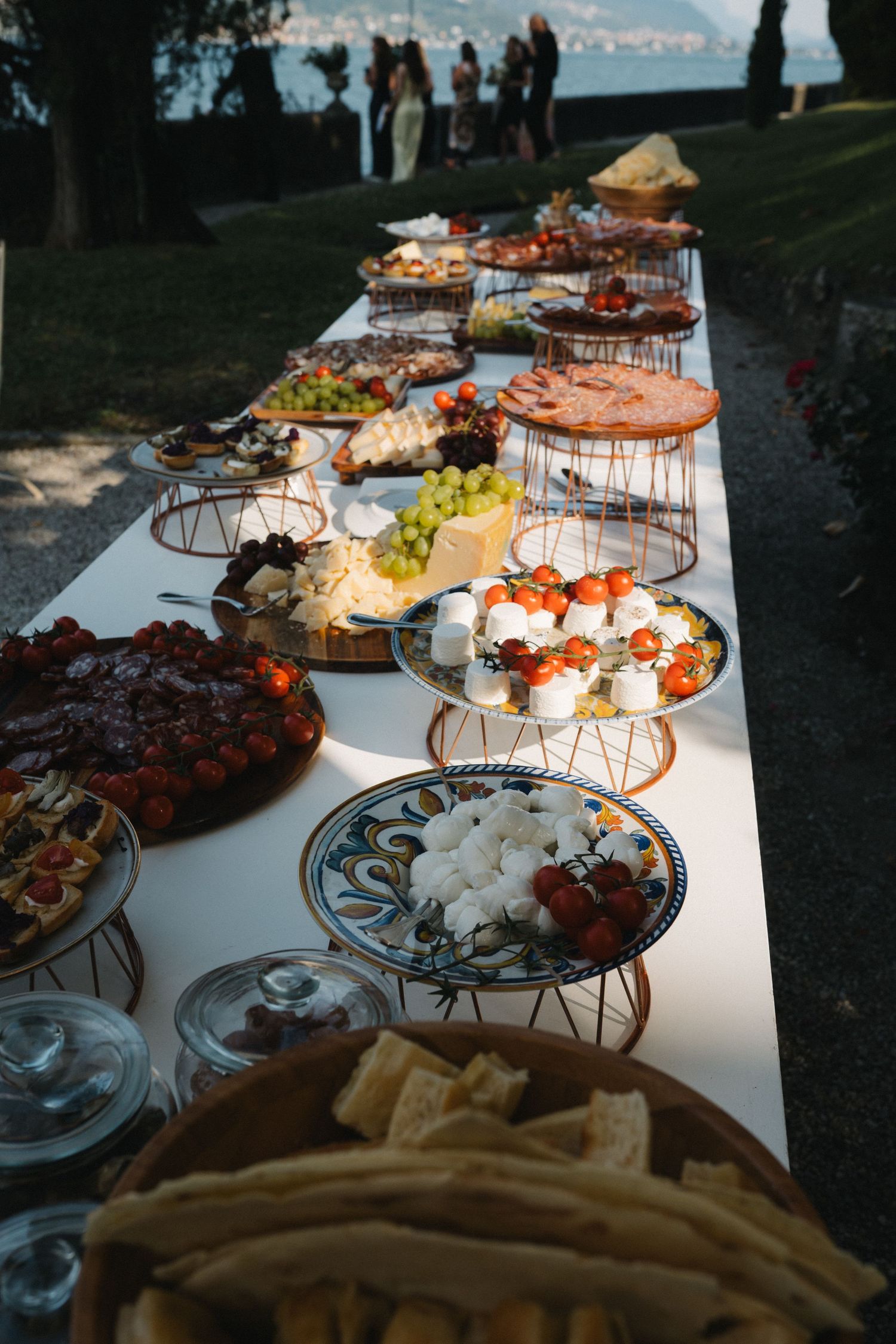 Elegant outdoor buffet table with charcuterie, fresh fruits, cheeses, and appetizers arranged on tiered stands by a lake.