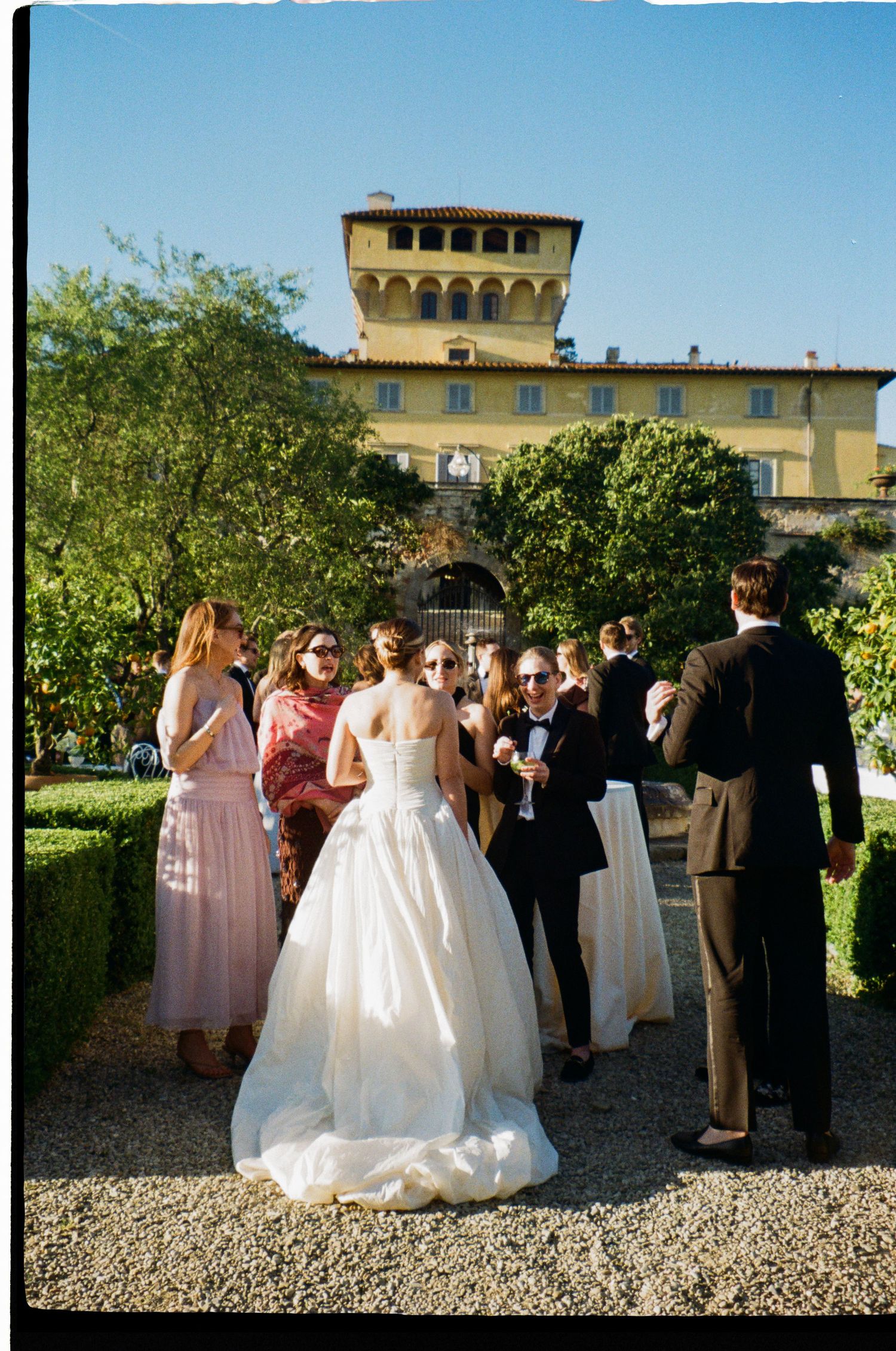 Bride in white ball gown mingles with guests outside a yellow Italian villa during cocktail hour.