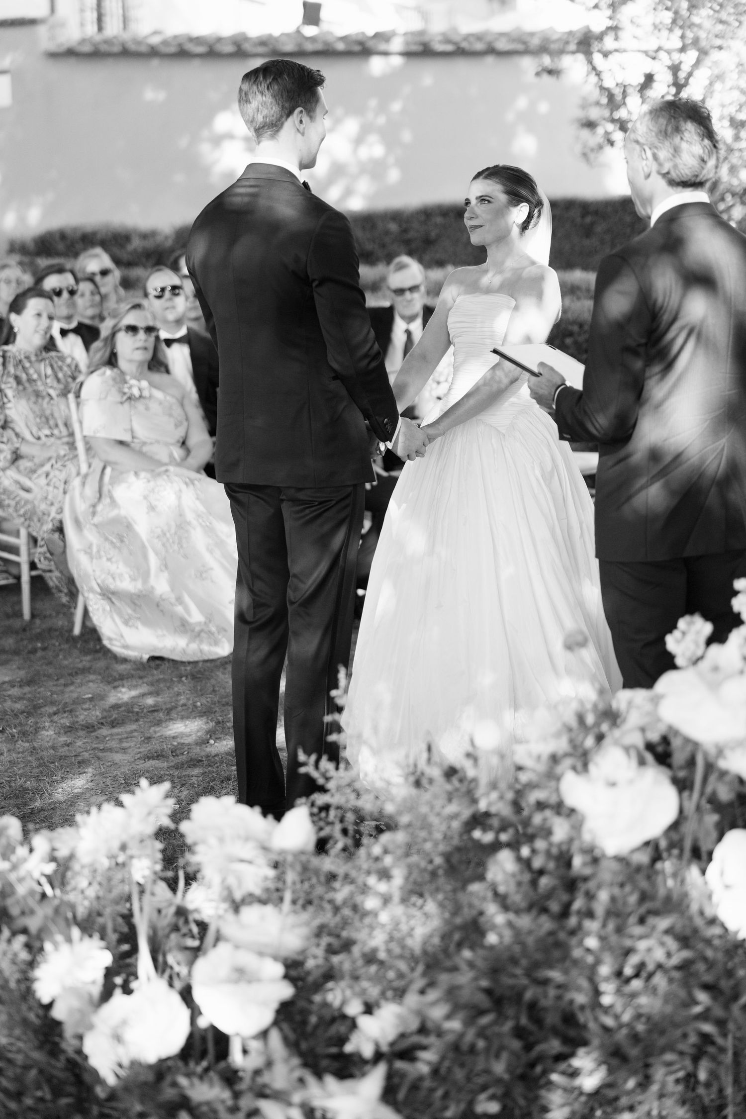 Black and white outdoor wedding ceremony with bride in off-shoulder gown holding hands with groom, guests seated behind.