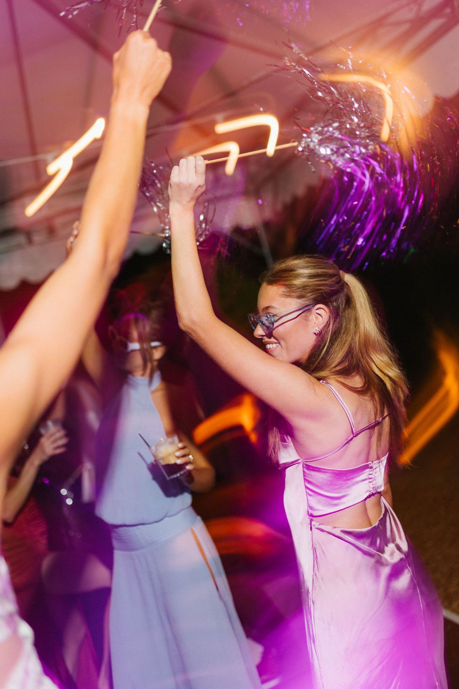 Women dancing at a party with sparklers and purple lights in elegant dresses.