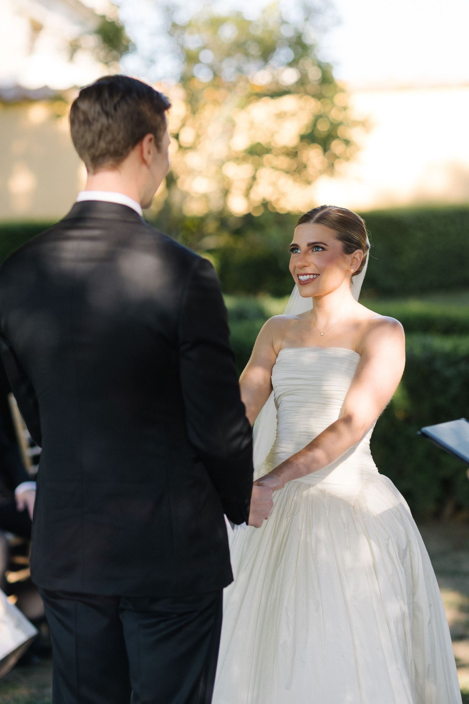 Smiling bride in strapless white tulle gown holds hands with groom in black suit during outdoor wedding ceremony.