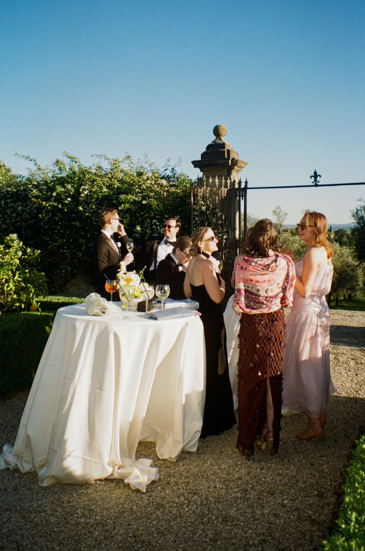 Wedding guests mingle at cocktail hour around draped table in sunlit Italian garden.