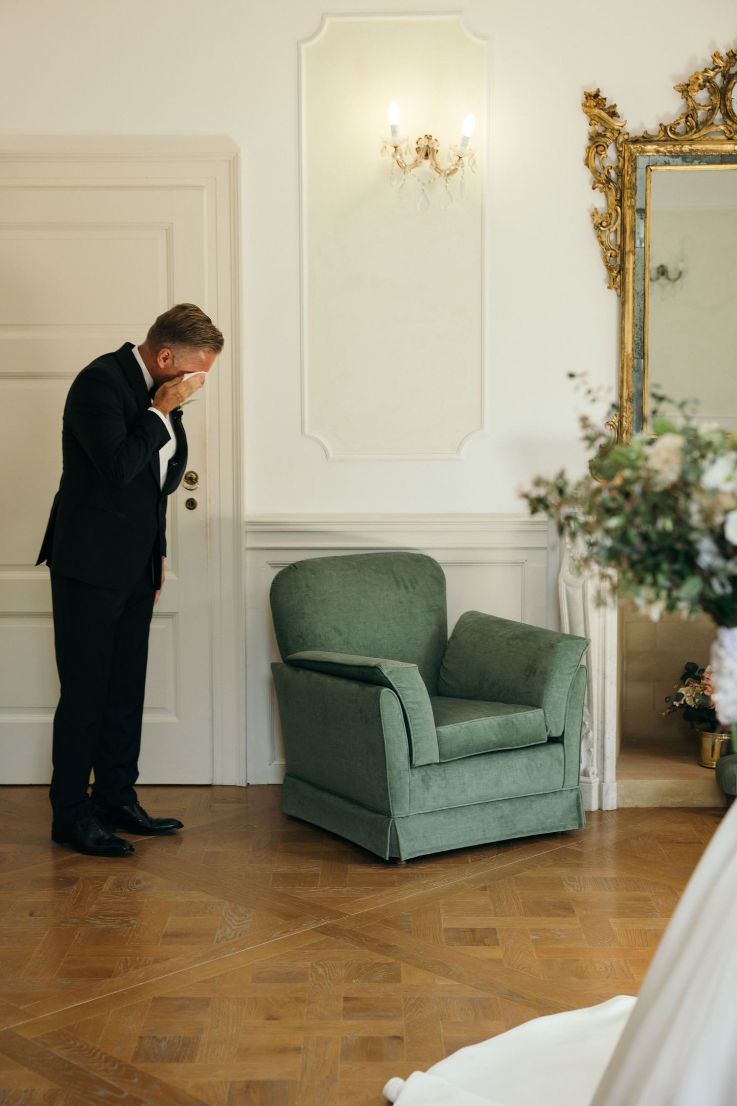 Groom in black tuxedo bowing his head emotionally beside a green velvet armchair in an elegant white room.