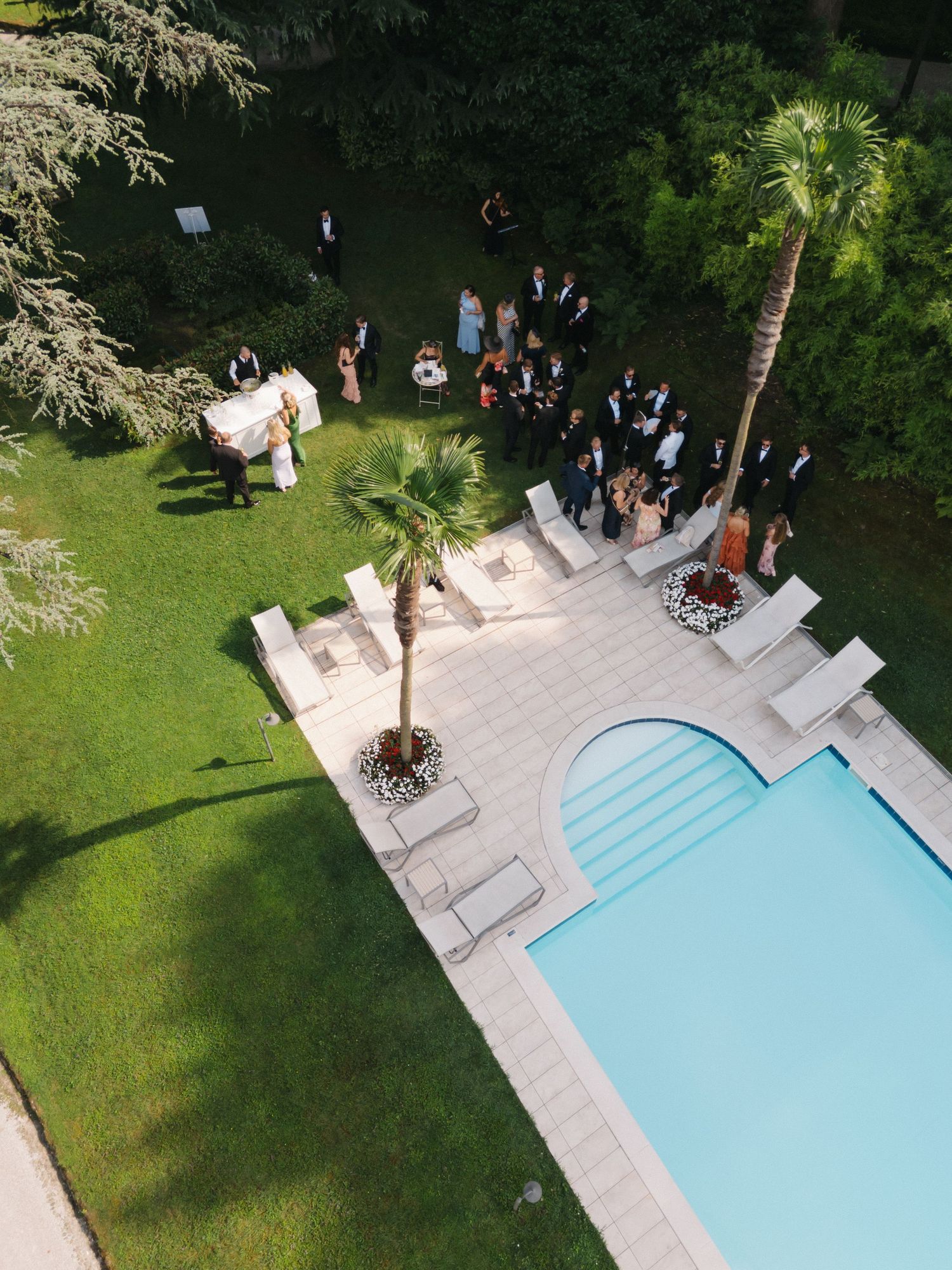 Aerial view of an elegant outdoor wedding reception beside a pool, with guests gathered on a terrace surrounded by lush greenery.