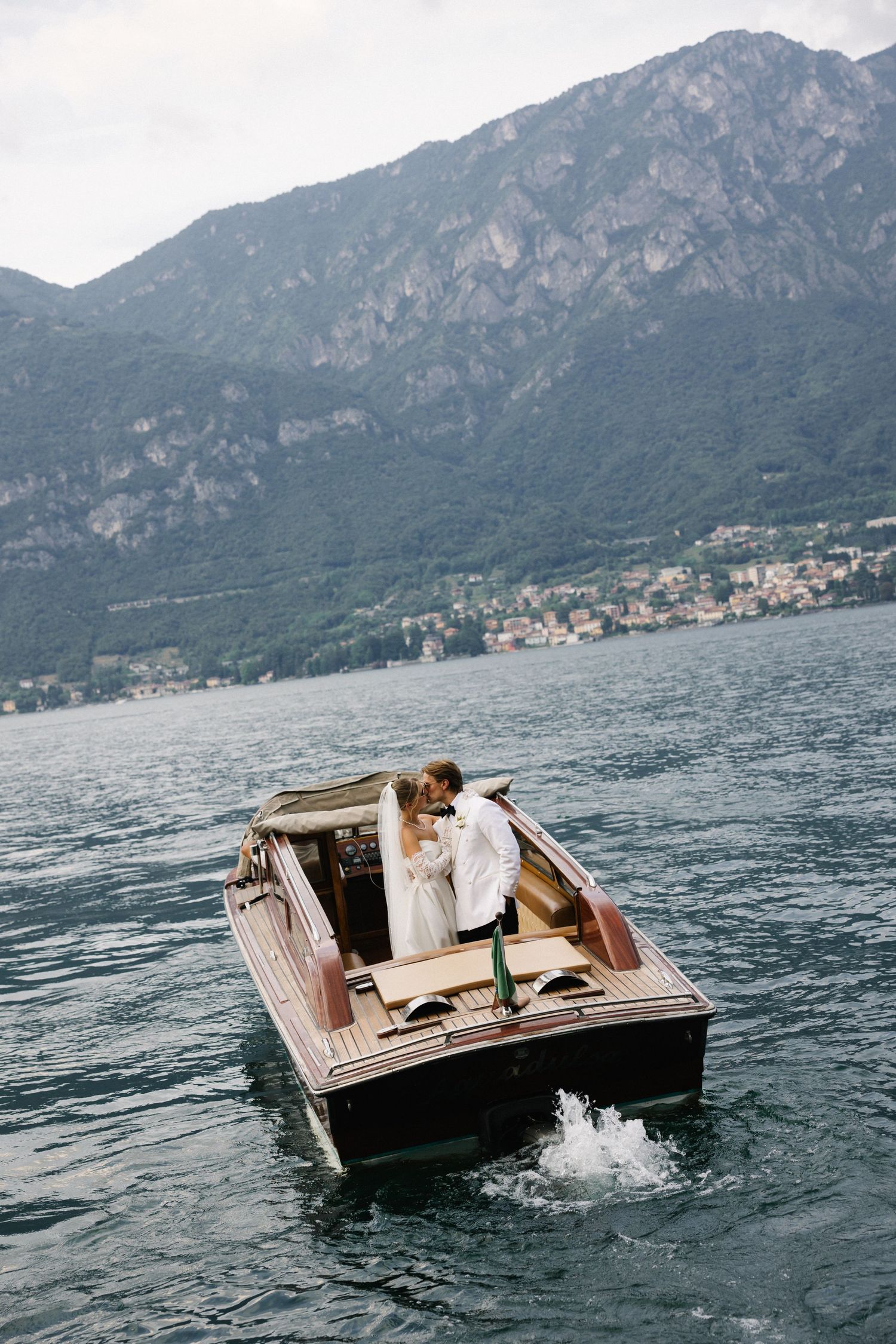 Couple kissing in a wooden boat on a lake with mountains and a hillside town in the background.