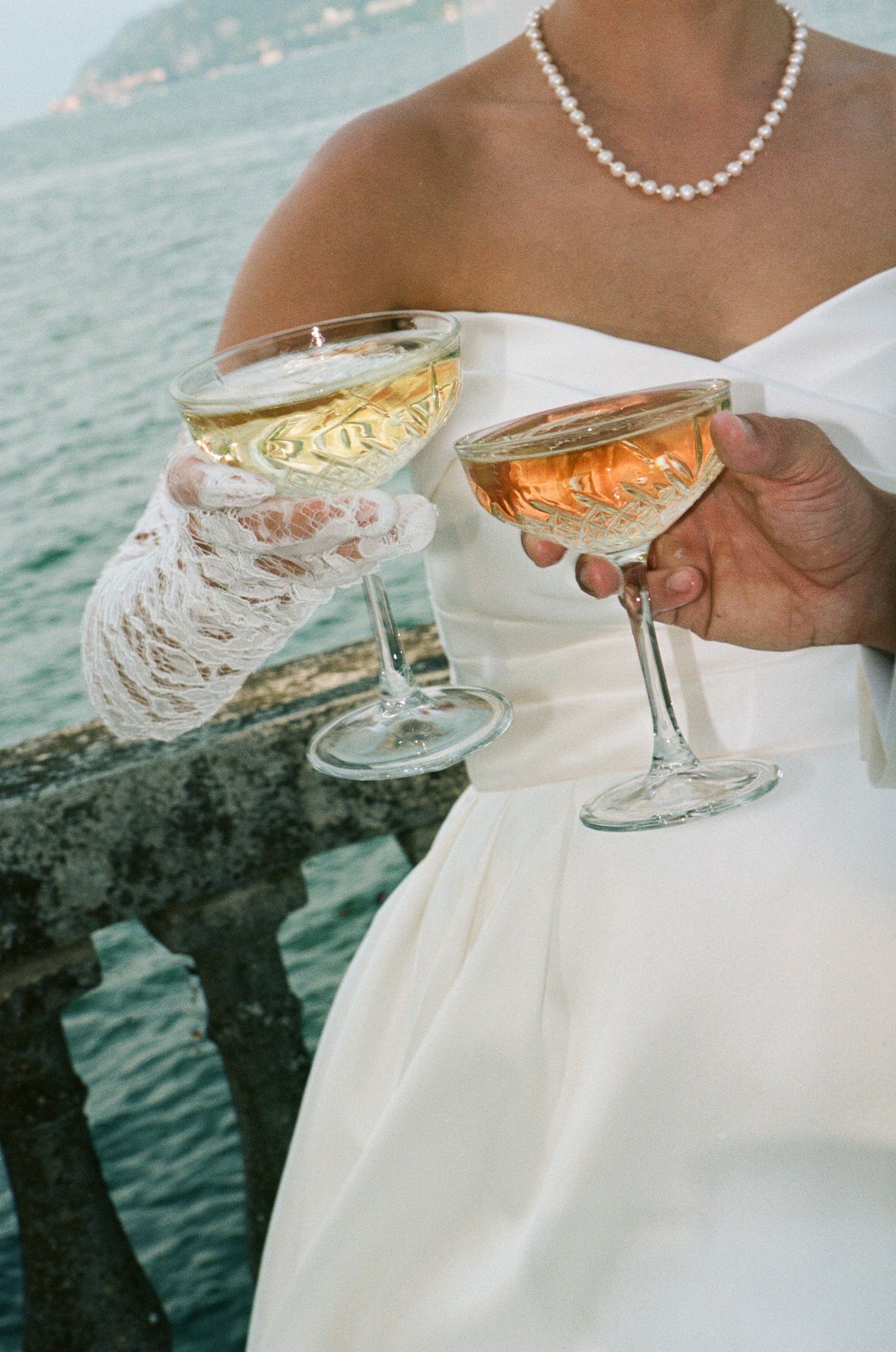 Bride in white strapless gown and pearl necklace holding two champagne coupe glasses near waterfront pier.