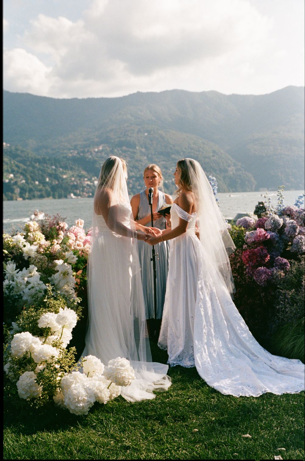 Two brides in white gowns and veils exchanging vows at floral altar overlooking Lake Como mountains in Italy.
