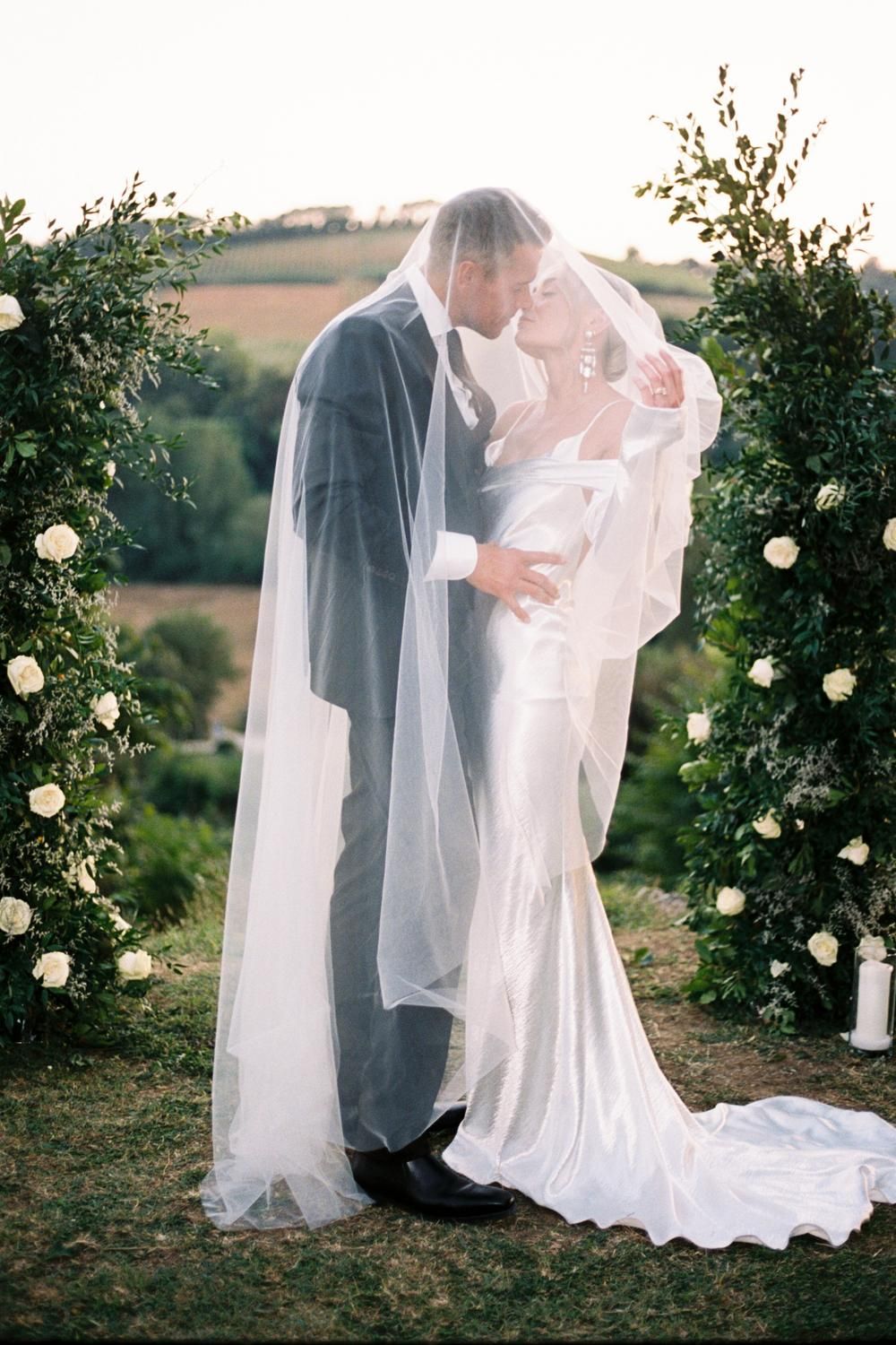 Romantic couple kissing under bridal veil at outdoor wedding ceremony with floral arch.