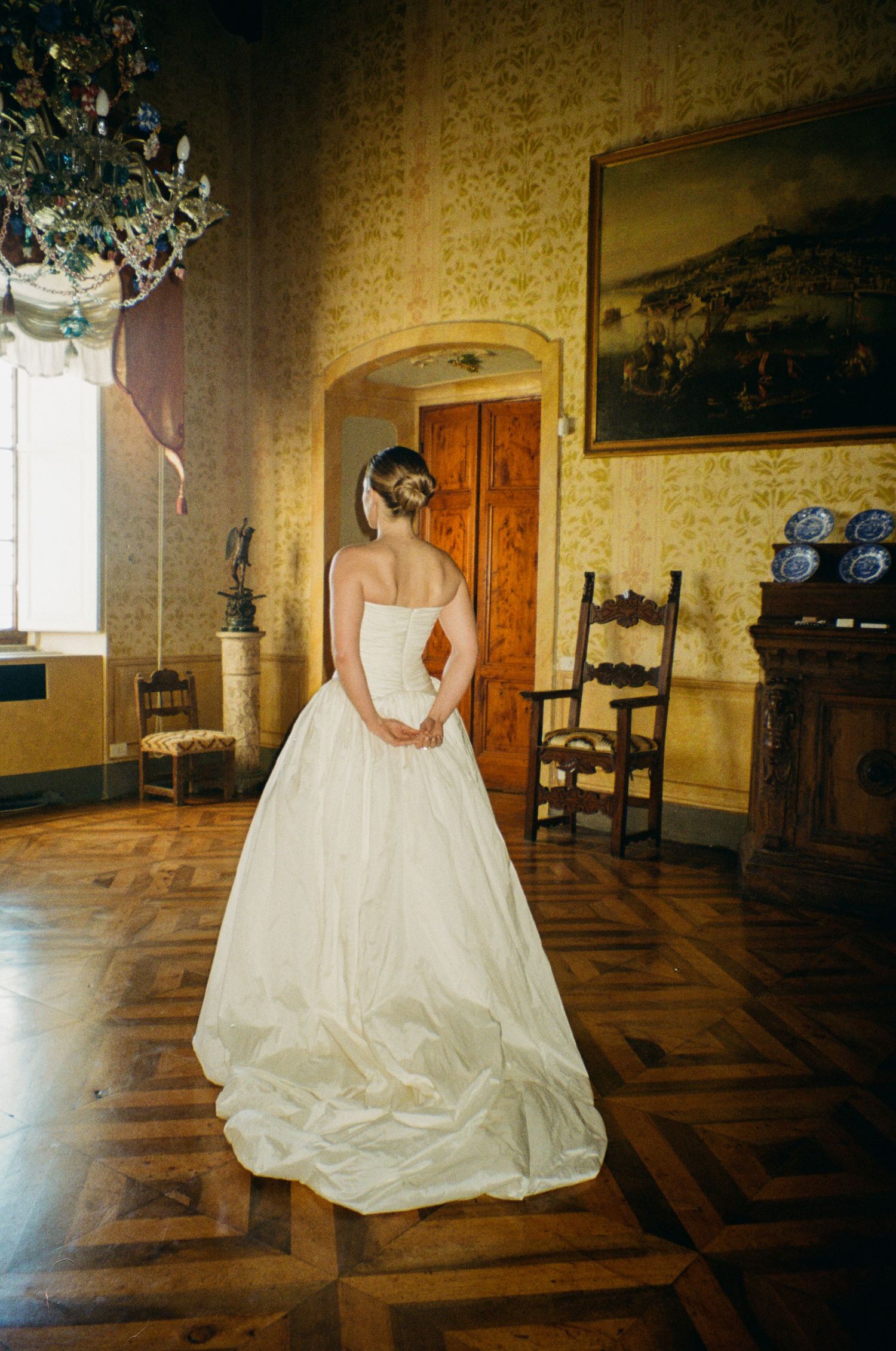 Bride in strapless white ball gown stands in elegant villa room with parquet floors and chandelier.