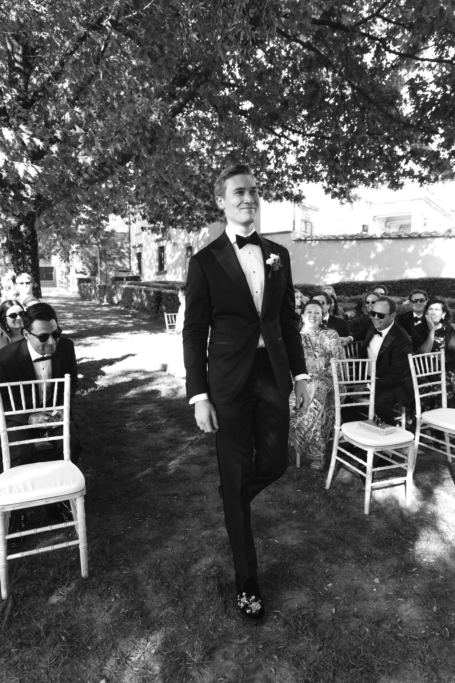 Groom in black tuxedo walks down outdoor aisle lined with white chairs at garden wedding ceremony.