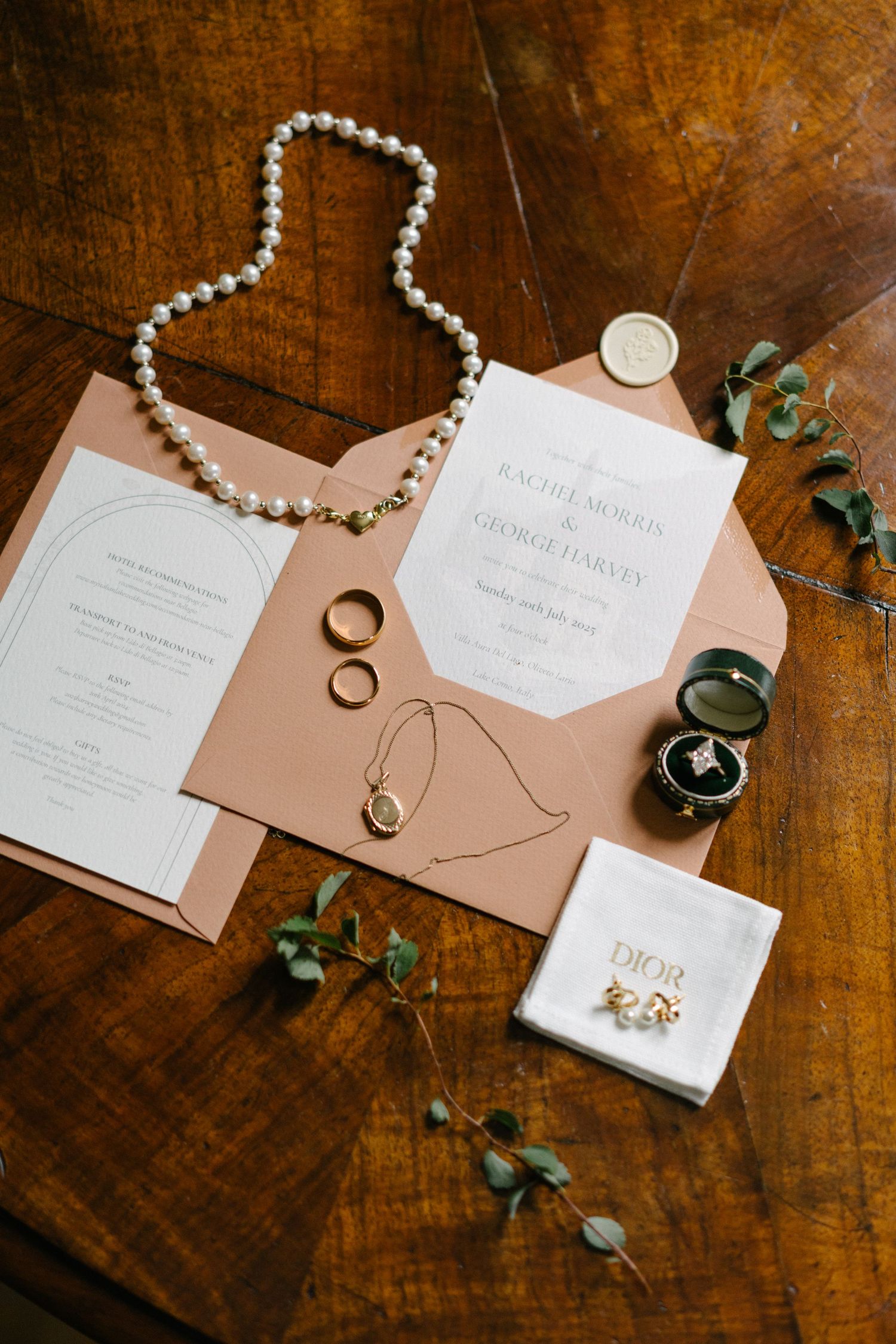 Elegant wedding flat lay with pearl necklace, gold rings, Dior earrings, invitation suite, and greenery on wood table.