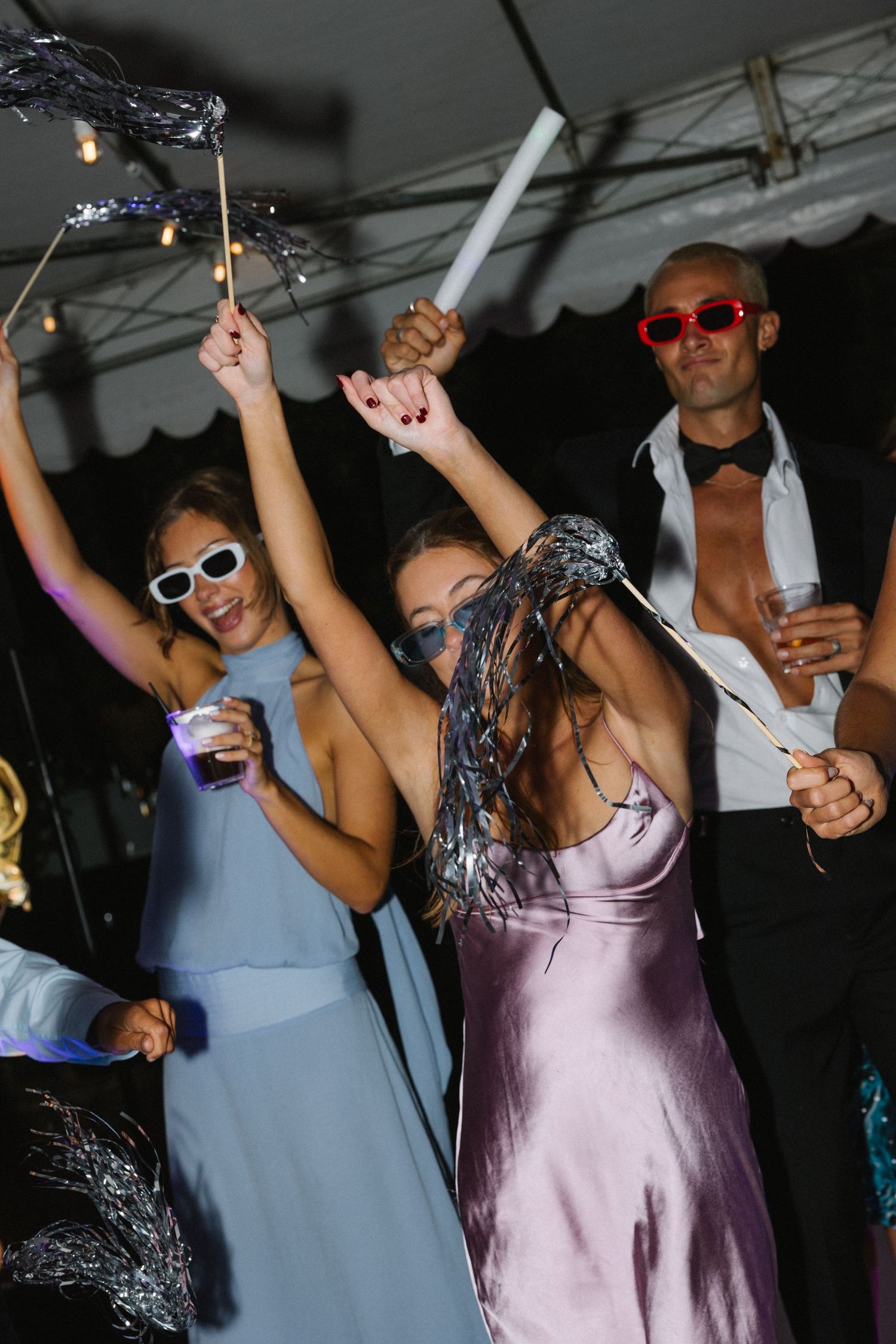 People dancing at a party in formal attire, wearing sunglasses and holding sparklers under a tent.