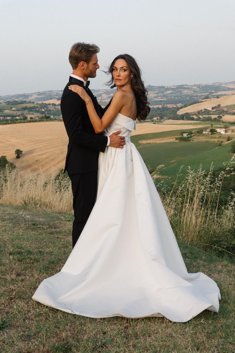 Bride in white strapless gown and groom in black tuxedo embrace on a hilltop with scenic Tuscan countryside views.