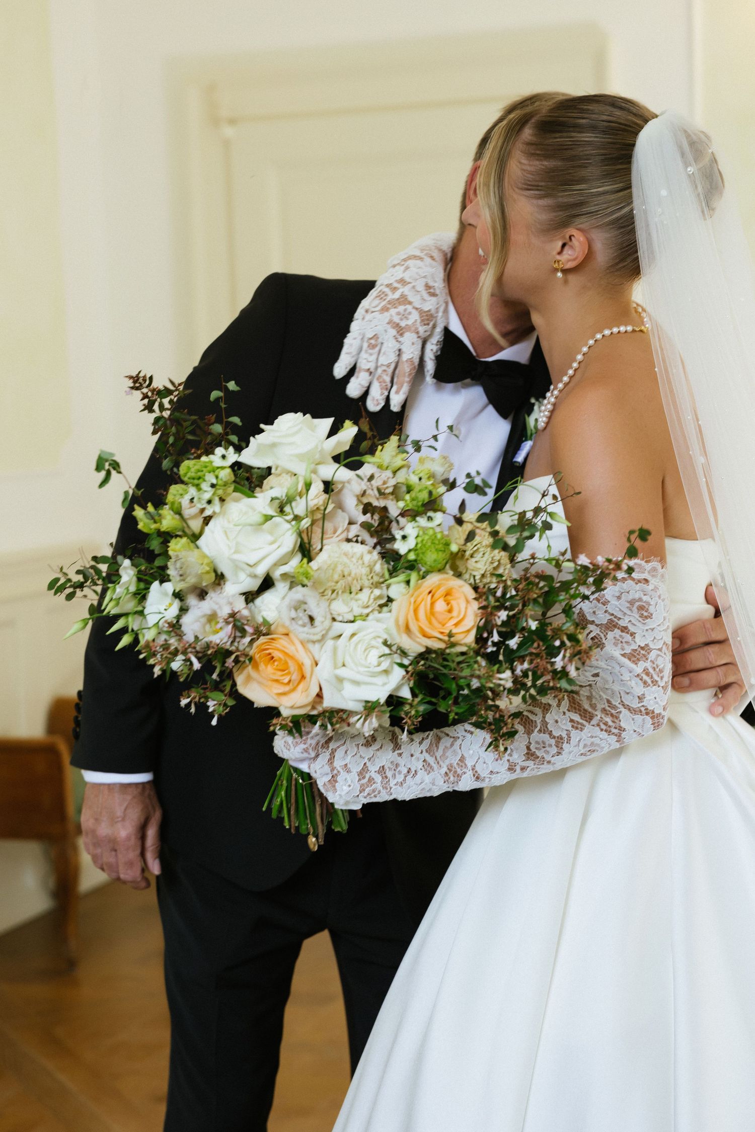 Bride and groom share a kiss, bride holding a lush bouquet of white and peach roses, wearing lace gloves and veil.