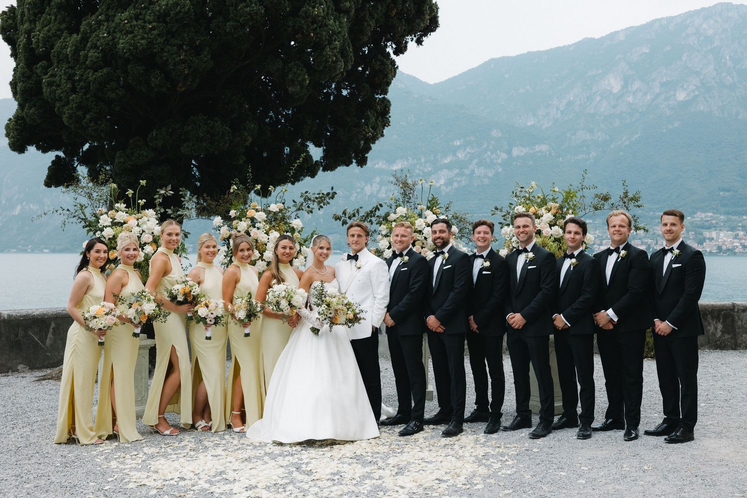 Wedding party poses lakeside at Lake Como, bridesmaids in gold dresses with bouquets beside groomsmen in black tuxedos.