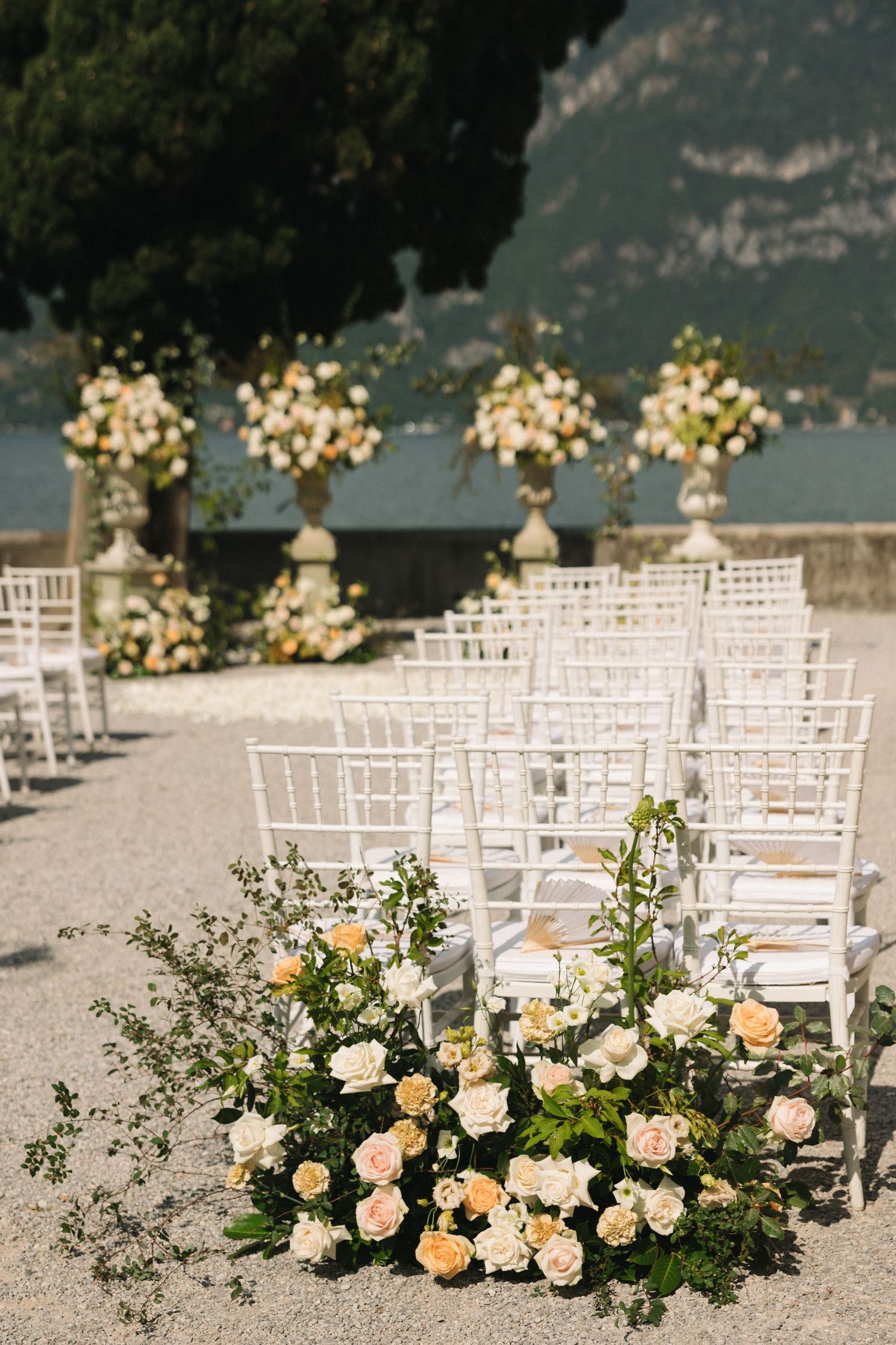 Elegant outdoor wedding ceremony with white chiavari chairs, peach and white rose arrangements, and stone urns by a lake.