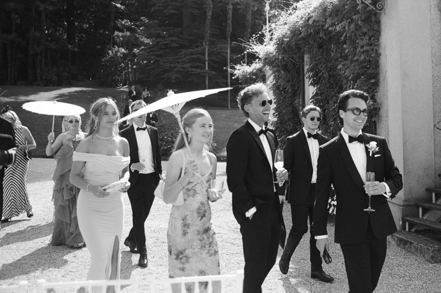 Black and white photo of elegantly dressed wedding guests in tuxedos and gowns walking outdoors at a formal event.