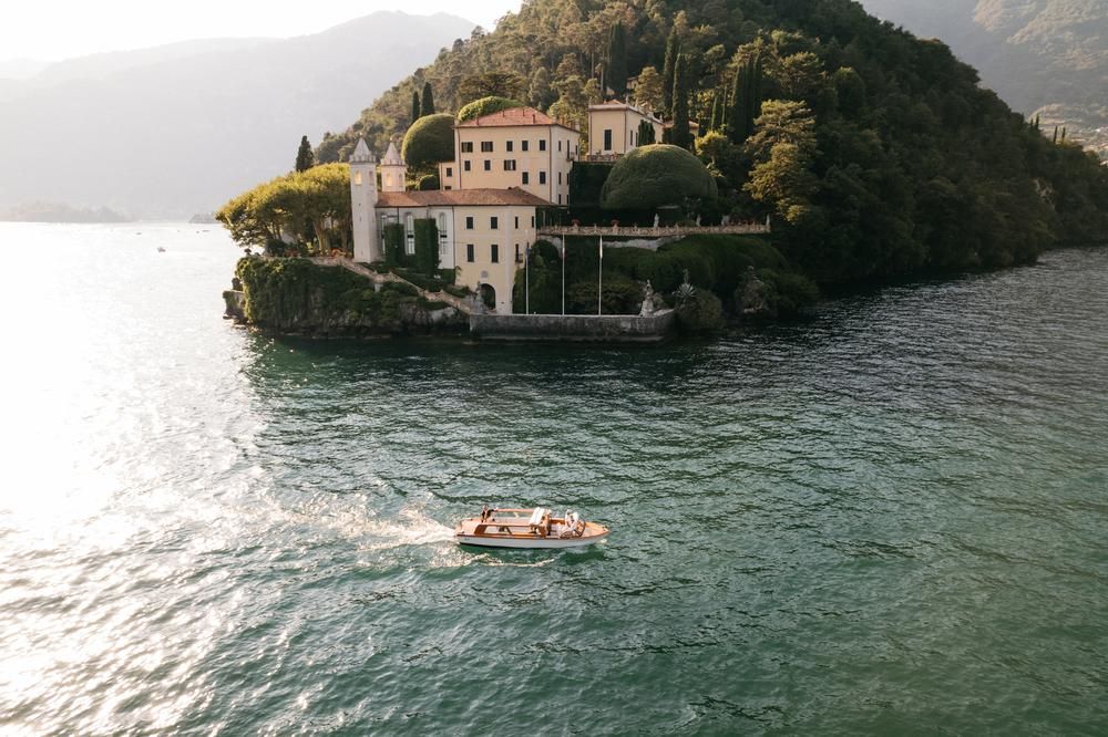 Aerial view of a classic wooden boat passing a stunning Italian villa on the shores of Lake Como.